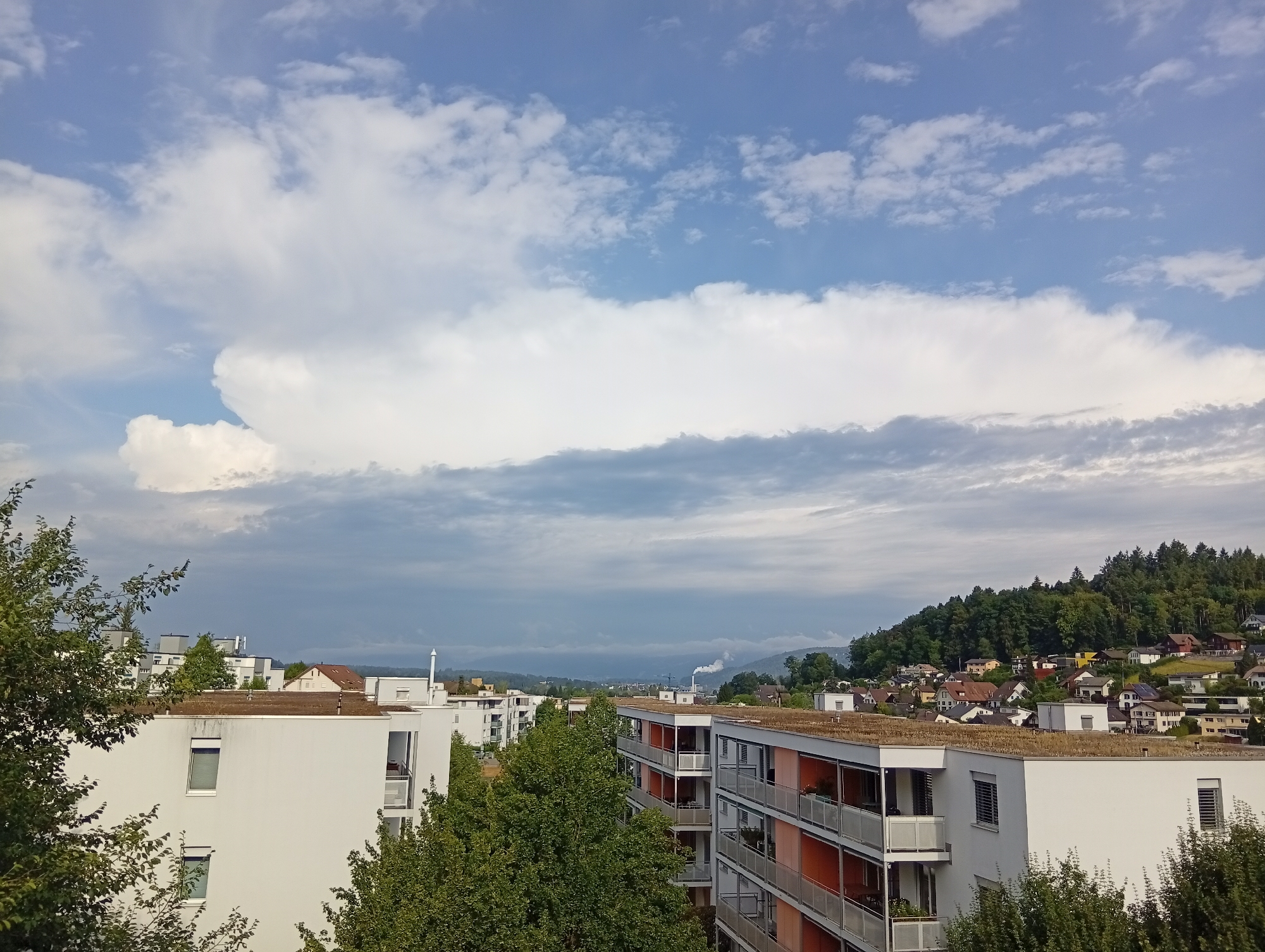 The image depicts a view of the city of Zofingen, Switzerland, from an elevated perspective, likely a residential building. In the foreground, modern white apartment buildings with balconies are visible, some featuring orange accents. Lush green trees partially obscure the buildings, suggesting a pleasant season, possibly summer or early autumn. Beyond the apartment complexes, the landscape transitions to a hillside densely covered with trees, dotted with houses, indicating a suburban or semi-rural area bordering the city. The sky is a prominent feature, filled with a mix of white cumulus clouds and darker, more ominous clouds, hinting at potential changing weather conditions. In the distance, a hazy view of more distant hills and industrial structures, possibly a factory with a smokestack emitting white smoke, can be observed. The overall scene conveys a sense of urban development integrated with natural landscape.