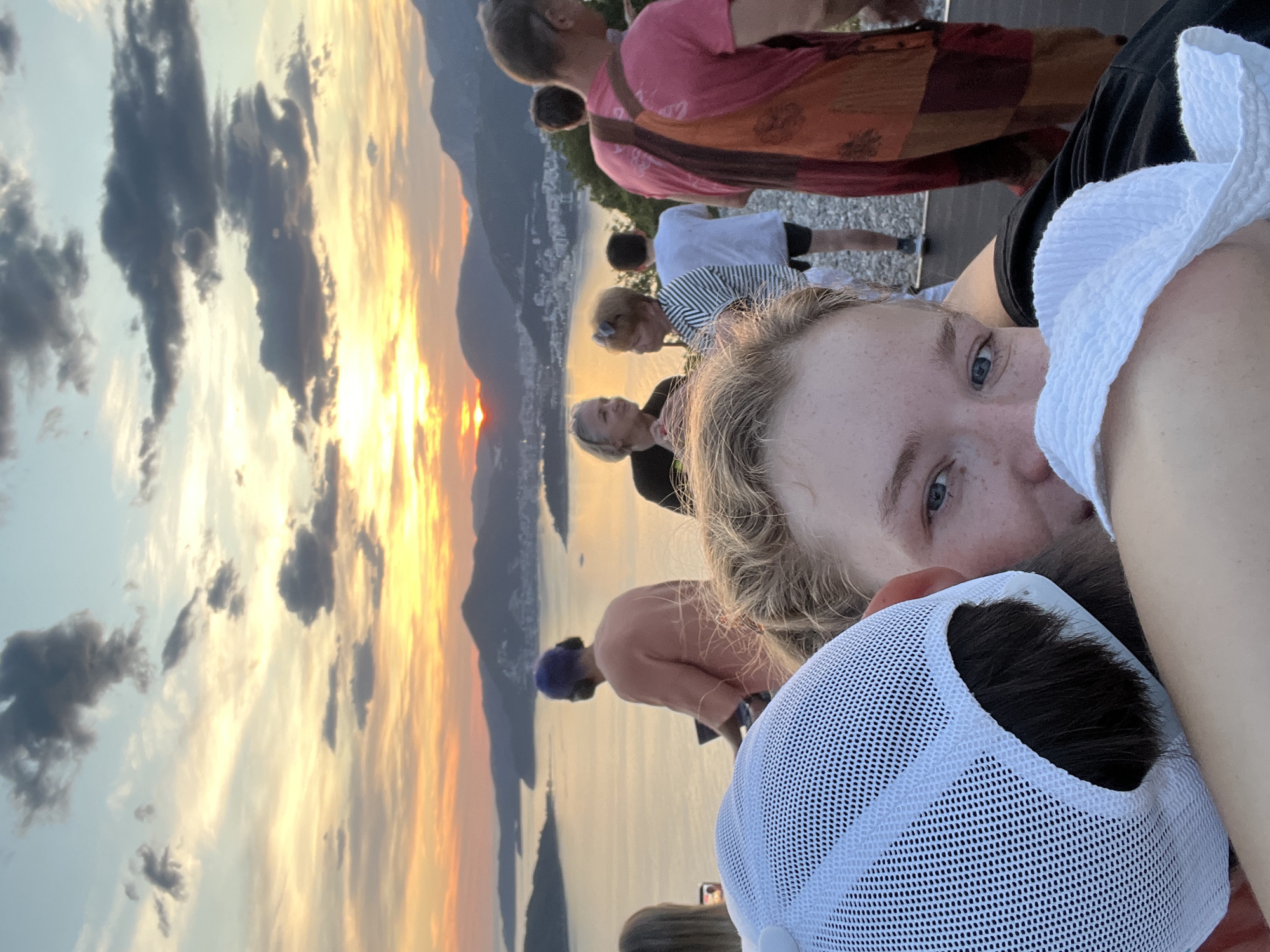 A close-up selfie captured at sunset, possibly in or near Budva, Montenegro, is dominated by two people: a blonde-haired individual with blue eyes, and another person wearing a white mesh baseball cap. Others are visible in the background, including some wearing a pink shawl and a striped shirt. In the distance, there is a mountain range with a sunset. The lighting indicates dusk or dawn. The background includes what looks to be a sea, with a city visible at the water’s edge. The selfie seems to be taken from a high vantage point. The people in the background appear to be tourists, and the overall scene suggests a popular location for sunset viewing.