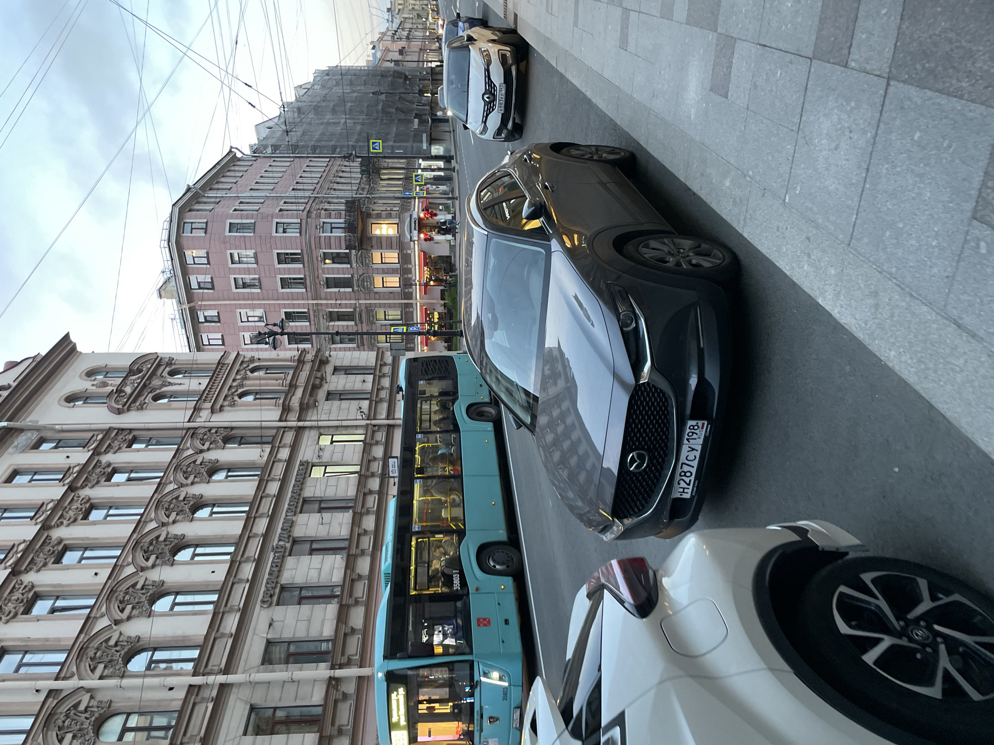 The image shows a street scene in Saint Petersburg, Russia, with parked cars and a public bus. A gray Mazda sedan with license plate "H287CY 198" is prominently featured. To its right is a partially visible white car. Behind the Mazda is a light blue bus. Further down the street, other vehicles are visible. Buildings line the street, with ornate architectural details. Overhead electrical wires crisscross the sky. The overall lighting suggests an overcast day.