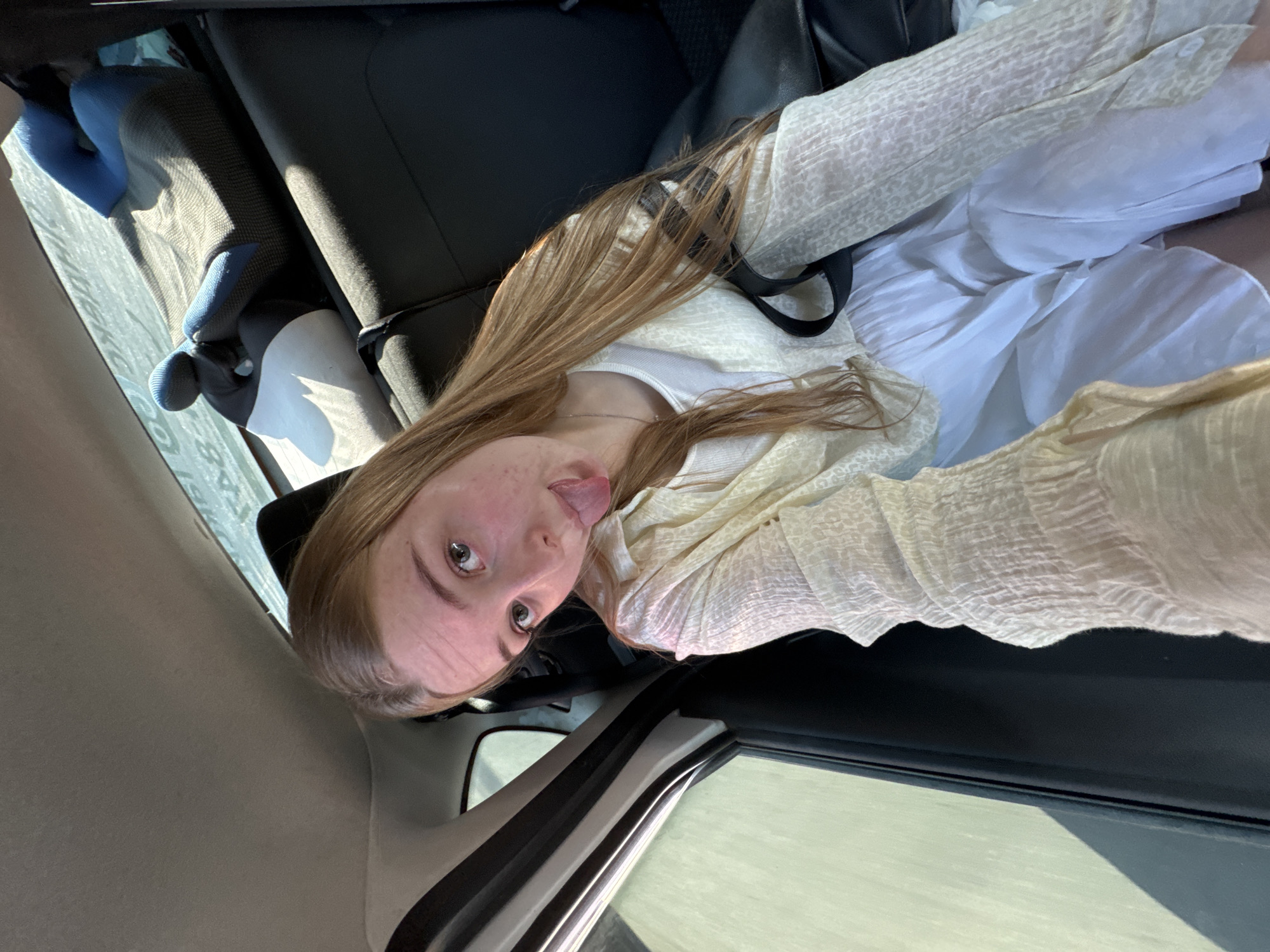 A young woman is taking a selfie inside a car. She is sticking her tongue out and looking at the camera. She is wearing a white top and a cream-colored cardigan. She has long, light brown hair. The interior of the car is dark, with black leather seats. There is a car seat in the back of the car, next to the woman. The car appears to be stationary as there is no indication of motion blur. The location is unknown, but it is claimed to be Chaikovsky, Russia.