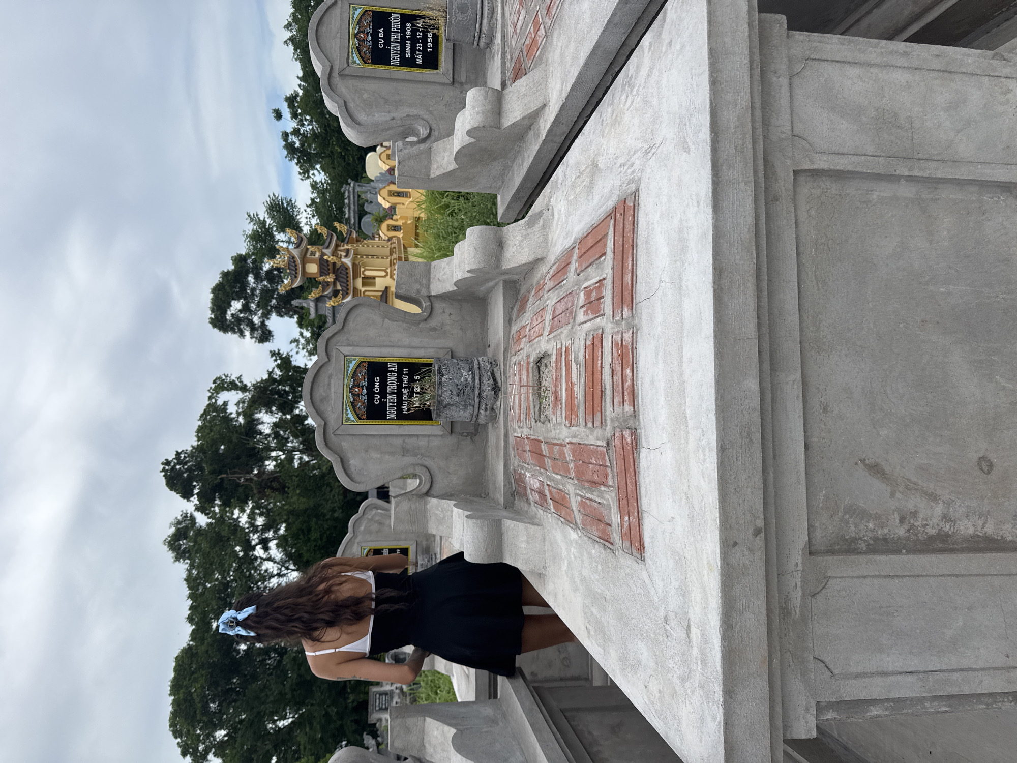 In a cemetery in Hoa Lư Ward, Vietnam, a woman with long, dark, curly hair, wearing a black dress and a white bra with a blue bow in her hair, stands facing away from the camera. She is positioned between two graves. The graves are made of concrete and feature engraved stone tablets with Vietnamese text. One tablet reads "CÙ ÔNG NGUYỄN TRONG AN SINH THỨ 11 MẤT 23 - 12 - AL" and the other reads "CÙ BÀ NGUYỄN THỊ PHƯƠNG SINH 1908 MẤT 23 - 12 - 1956". The graves are topped with red bricks arranged in a pattern, and some are cracked. In the background, there are trees and elaborate, ornate structures, possibly other graves or temple-like buildings, also adorned with gold accents. The sky is overcast.