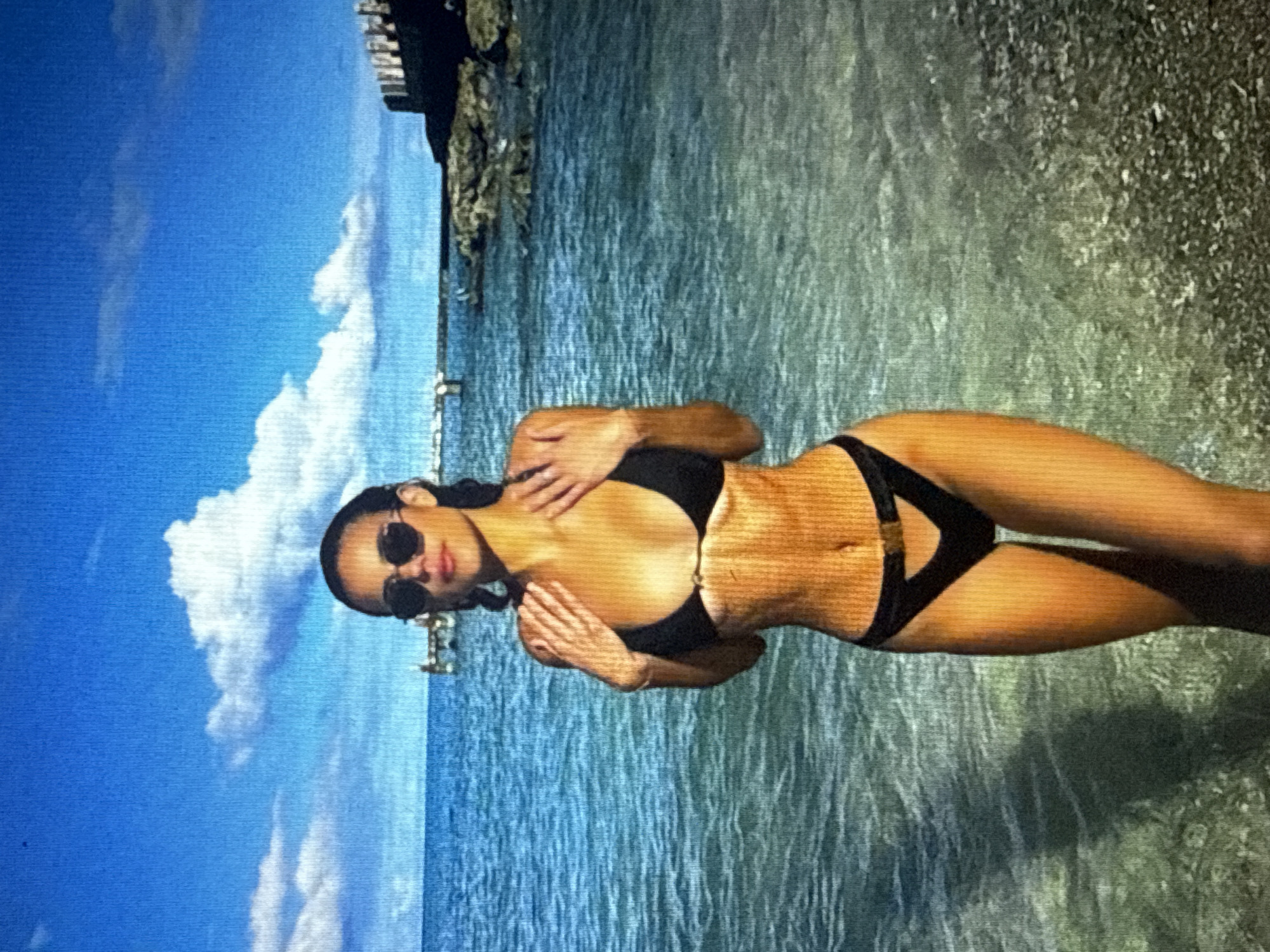 A woman wearing sunglasses and a black bikini stands in the shallow, clear blue water near a pebble beach. The ocean stretches out behind her, with a pier visible in the distance and a stone wall to the right. The sky above is a vibrant blue, dotted with white clouds. The woman has her hands on her chest, and her hair is dark and pulled back. The lighting suggests it is a sunny day.