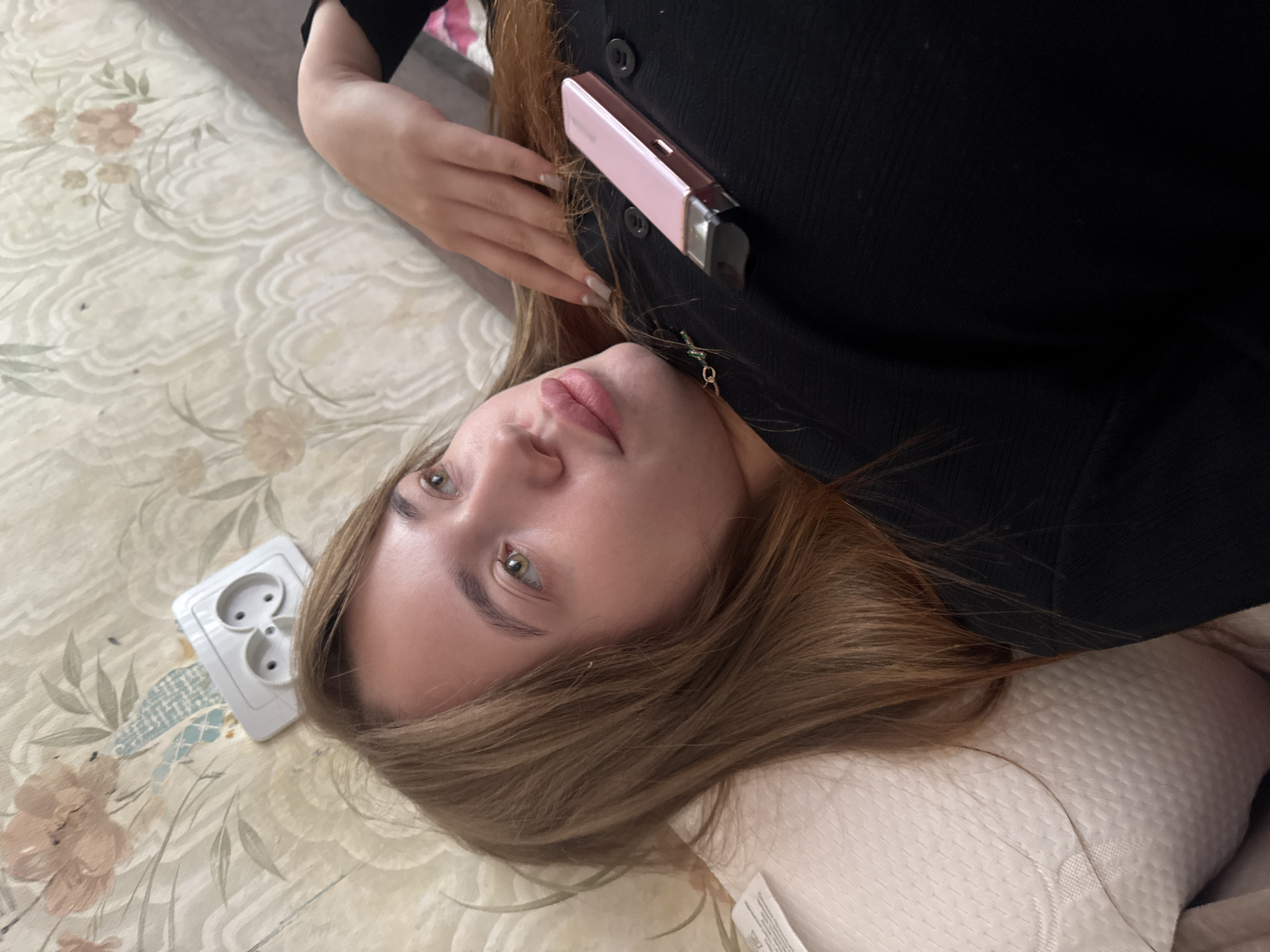 A woman with fair skin and auburn hair lies on her back on a bed with a floral-patterned bedspread. She is facing the camera with a slightly tilted head. The woman is wearing a dark-colored button-down shirt or dress. A small, light pink object, possibly a phone or cosmetic compact, is tucked into the neckline of her clothing. Her head rests on a white pillow.
A white European-style electrical outlet is visible on the bedspread near her head.
The available information does not allow the determination of the specific location in the city of Chaikovsky, Russia.