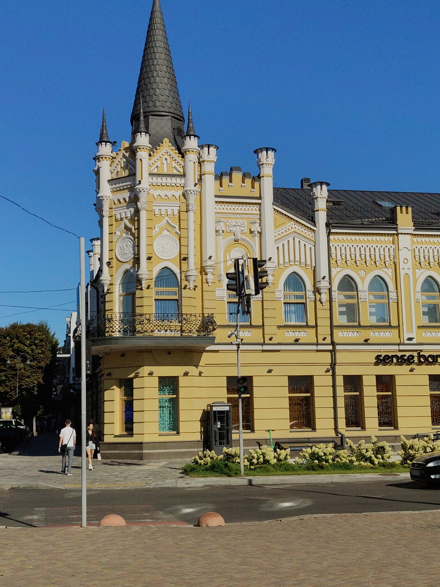 This is an outdoor, eye-level, full-shot photograph of a distinctive yellow building with an ornate, fairytale-like architectural style, located in Cherkasy, Ukraine. The building, which appears to be a former residential or public structure, features a prominent dark spire on its corner, flanked by smaller spires and decorative elements. The facade is adorned with white ornate trim, arched windows, and circular medallions. A balcony with intricate ironwork extends from the corner of the building. A sign that reads "sense bank" is visible on the lower section of the building. The building is situated on a city street with a crosswalk in the foreground. Two individuals are walking on the sidewalk. One is a male wearing a white t-shirt and grey pants, walking away from the camera, and the other is a female wearing a black jacket and denim shorts, also walking away from the camera. A traffic light stands to the left of the building, and a classic grey telephone booth is positioned near the corner. Lush green bushes with white flowers are planted in front of the building. The sky is a clear, vibrant blue, and the sun is casting shadows, indicating it is daytime. The foreground shows a paved sidewalk and a road. A black car is partially visible on the right side of the image.