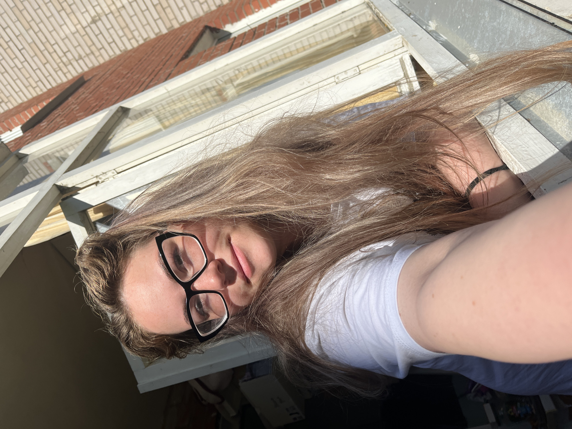 A young woman is taking a selfie in Chaikovsky, Russia. She is wearing glasses and a white t-shirt. Her long, brown hair is partially obscuring her face. She is either at an open window or on a balcony, as a white window frame is visible. Behind her is a brick building. The image is well-lit and in focus.