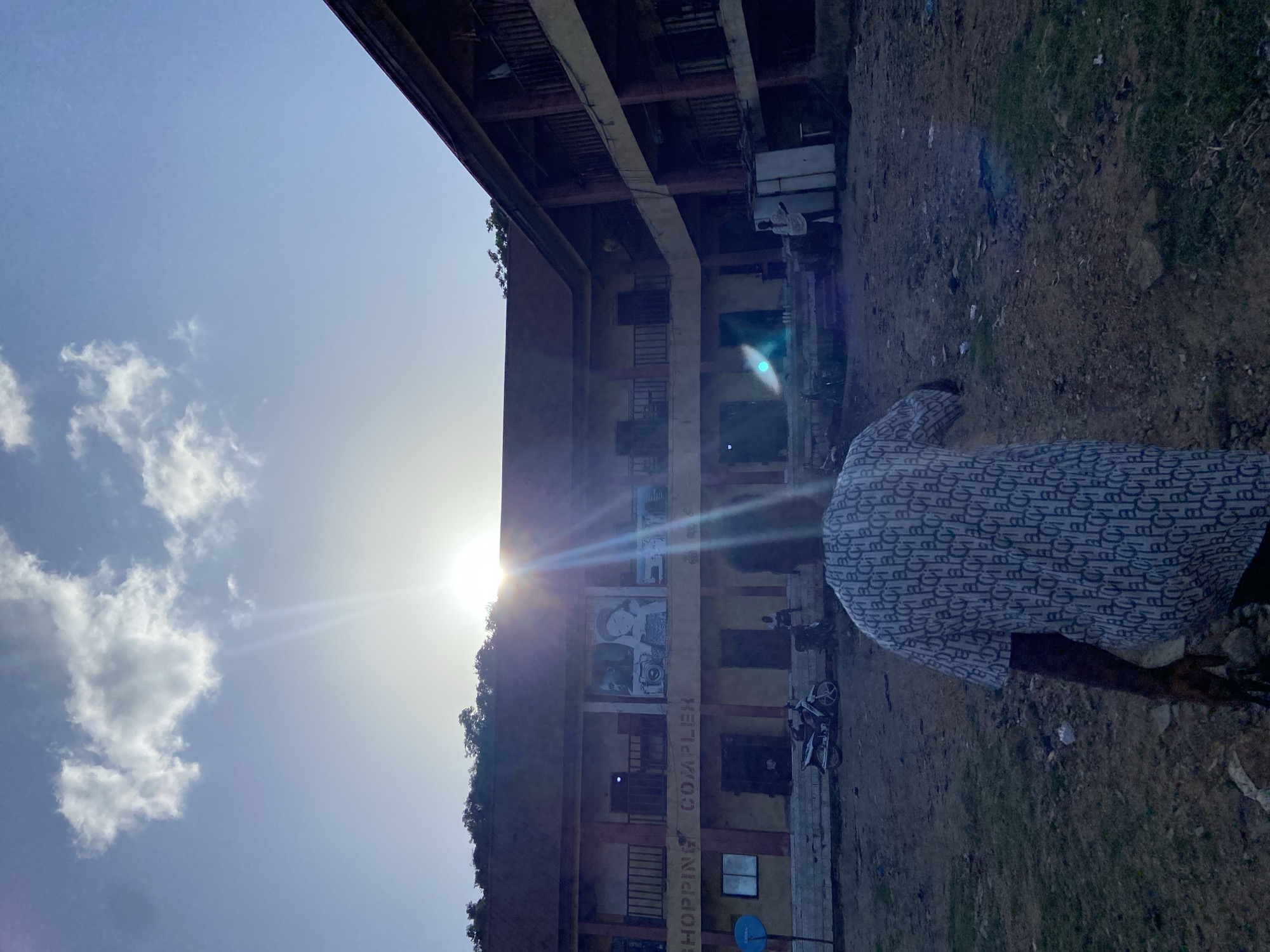 The photo is taken in Mararaba, Nigeria. It shows a multi-story building with "SHOPPING COMPLEX" painted on it. A figure in a patterned shirt is present, and several motorcycles are parked nearby. The photo is taken from a low angle, with the sun positioned behind the building. The sky is visible with some clouds. The surrounding area is dirt.