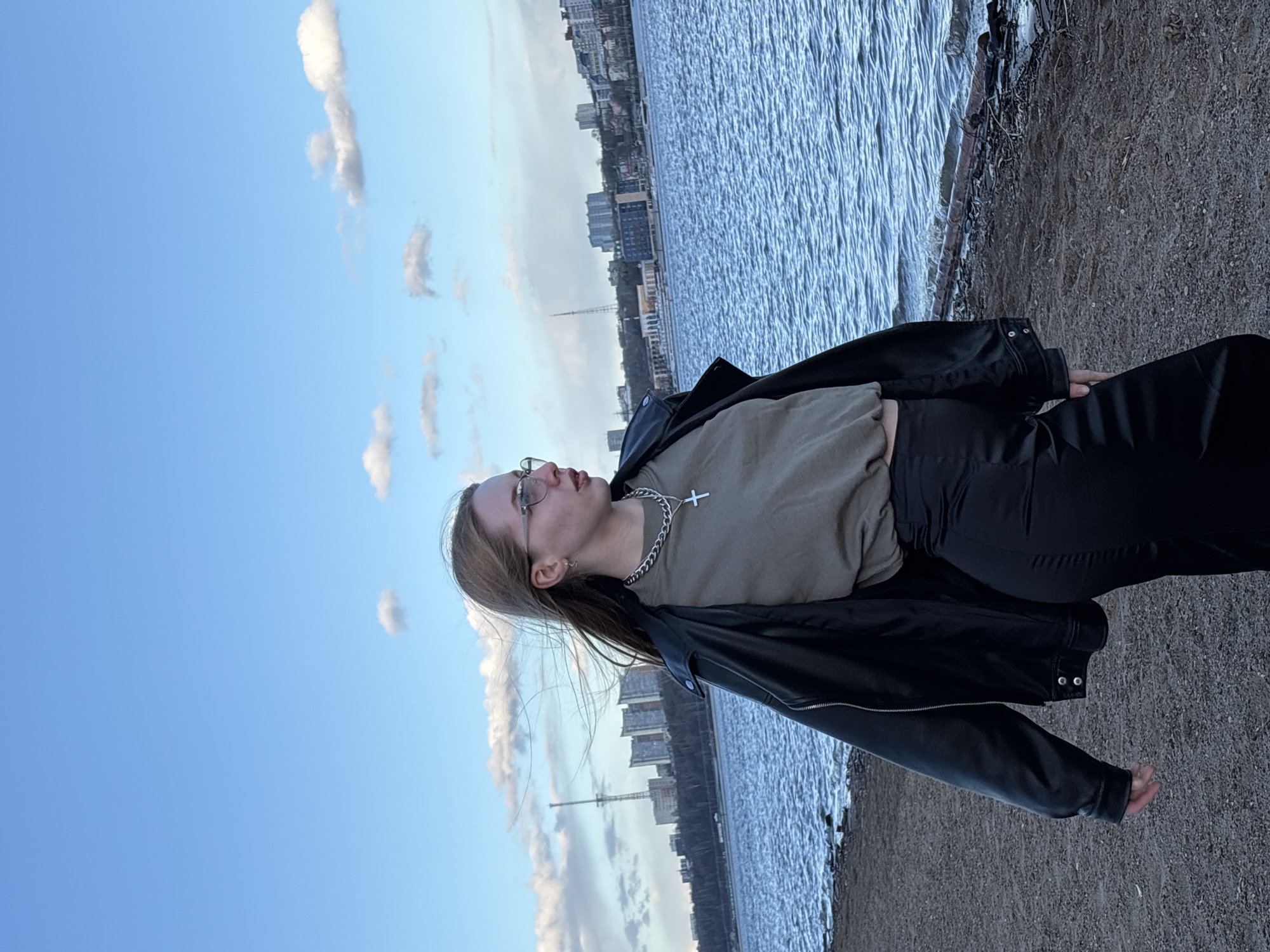 A portrait-oriented image captures a young person standing on a sandy or gravelly riverbank, gazing towards the horizon. The subject, likely female, is positioned on the right side of the frame, viewed in profile from the chest up. She has long, straight brown hair and wears clear-framed glasses. Her expression is contemplative as she looks upwards and to the left. She is dressed in a light olive green cropped t-shirt, a black leather-style jacket, and black pants. A distinctive silver chain necklace with a cross pendant is visible around her neck, along with small hoop earrings.
To the left of the person, a wide body of water, likely a river or large lake, dominates the midground. Its surface is gently rippled, reflecting the ambient light. Across the water, a distant cityscape forms the background, featuring an array of urban buildings and a prominent, tall slender structure, possibly a tower or mast. The sky above is a clear, bright blue with scattered, wispy white clouds. The lighting suggests late afternoon or early evening, creating a serene atmosphere. The scene depicts an urban waterfront setting, consistent with the described location of Perm, Russia, possibly along the Kama River. No visible text is present in the image.