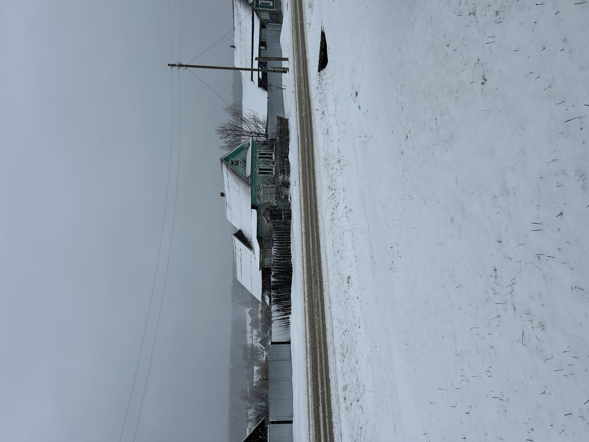 The image depicts a winter scene in Уральское, Russia. A snow-covered road runs parallel to a row of buildings, primarily houses. The buildings vary in size and construction; one prominent structure has a green roof and weathered wooden exterior. A wooden fence separates the buildings from the road. Power lines stretch across the sky. The sky is overcast and grey. The road surface shows tracks, presumably from vehicles. The ground is covered in a layer of snow with sparse vegetation poking through.