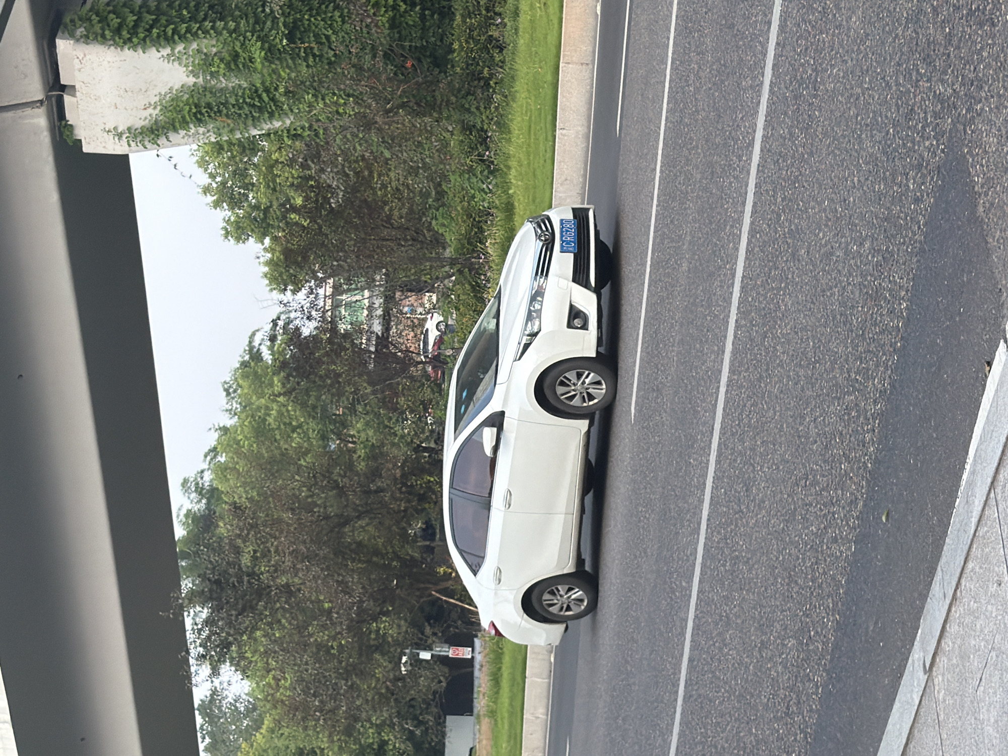 A white sedan is driving on a paved road, partially shaded by an overhead structure. The car's license plate reads "C RG280". Green trees and vegetation line the side of the road, and a building is visible in the background. The sky is overcast. The scene appears to be in Jiangjin, China.