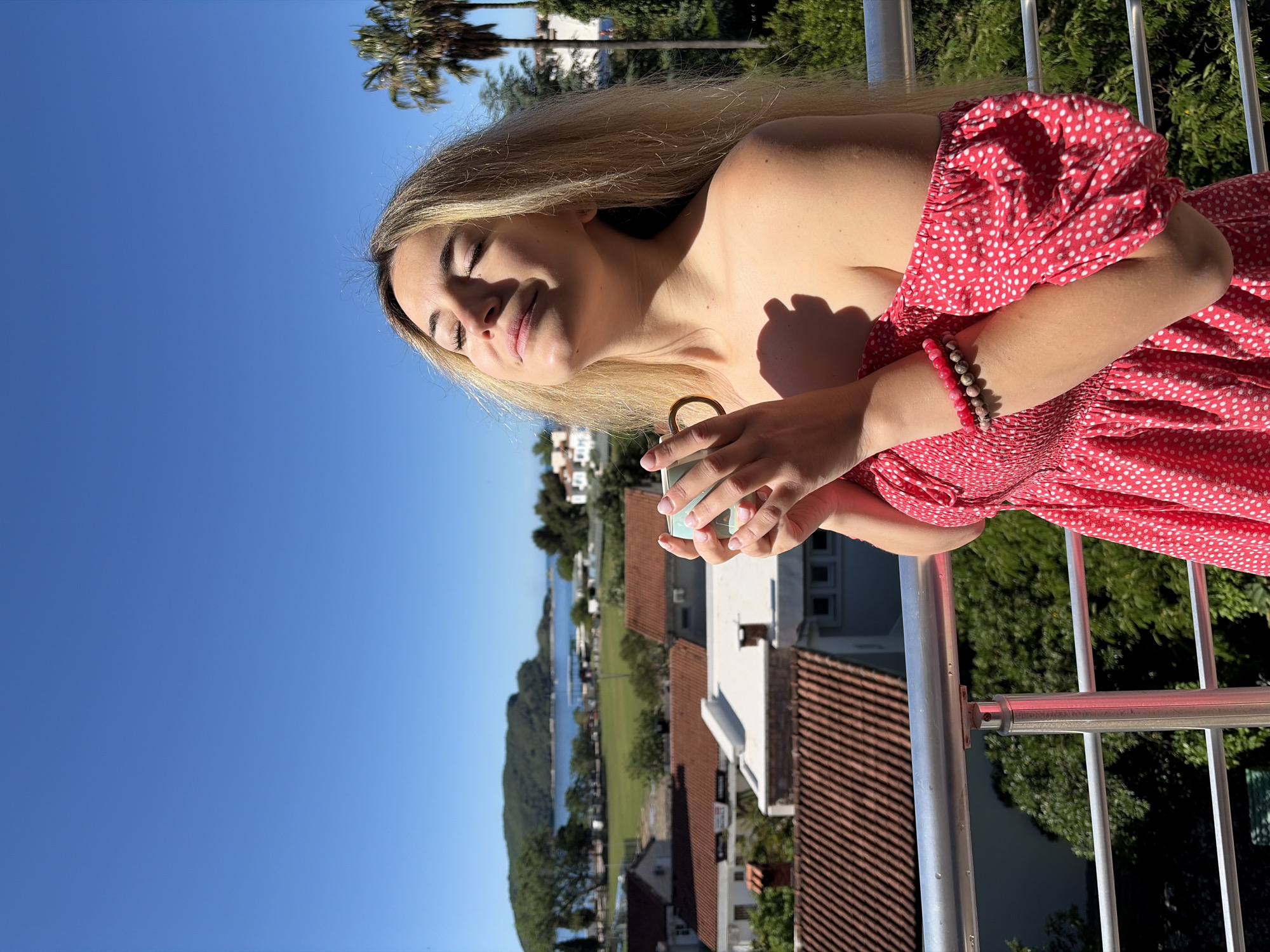 A woman with long blonde hair stands on a balcony in Budva, Montenegro, facing the sun with her eyes closed, wearing a red and white polka dot dress and beaded bracelets. She holds a small teal and white mug with a gold handle in both hands. Behind her, a residential area of Budva is visible, with red-tiled roofs and white buildings. Beyond the houses, there is a green field or park area, a body of water with mountains in the background, and a clear blue sky. Tall palm trees can be seen in the distance. The balcony railing is made of silver metal.