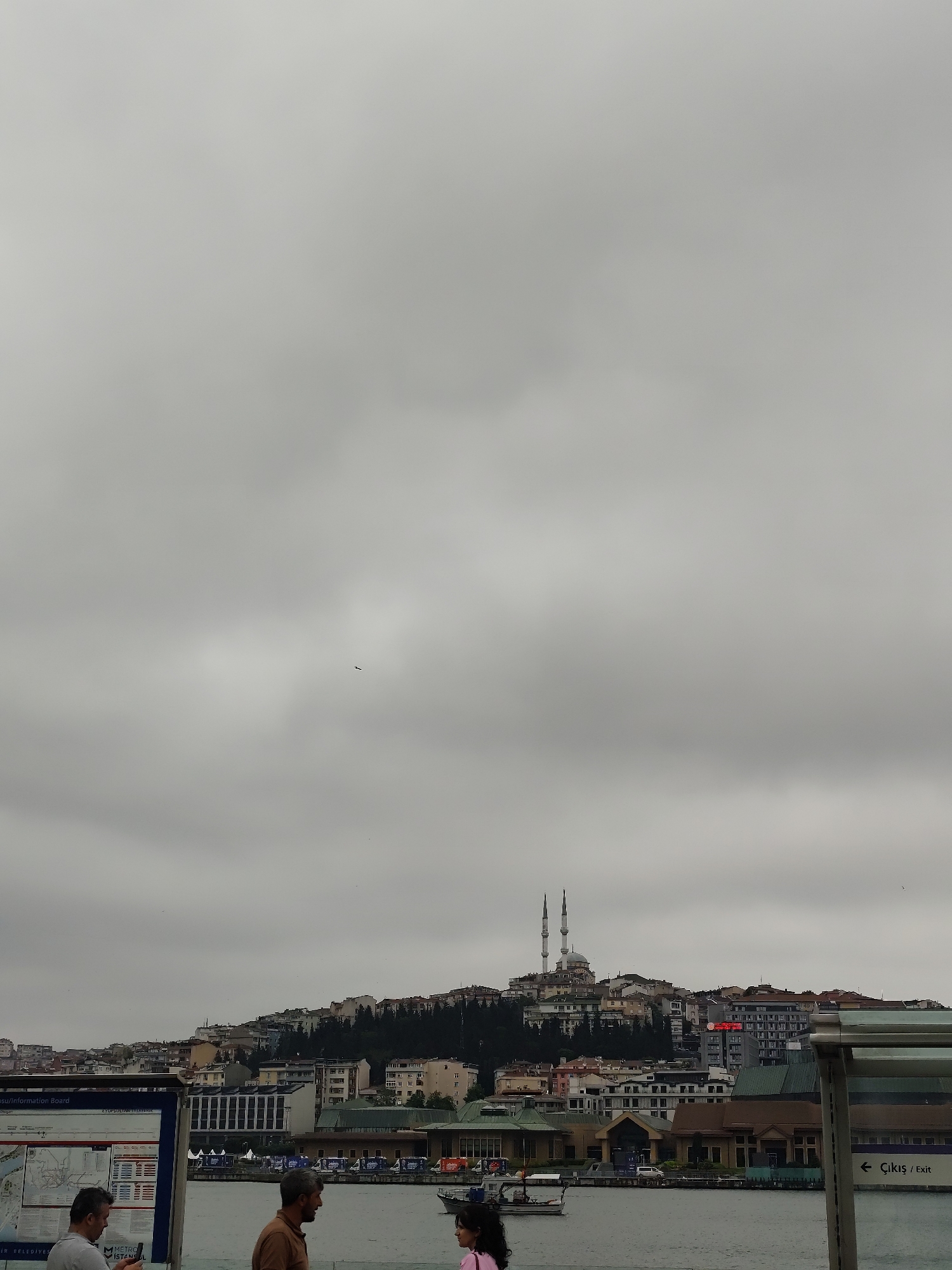 A low-angle, wide shot of a cityscape with a large body of water in the foreground, likely taken in Eyüpsultan, Turkey. The sky is overcast with a uniform gray cloud cover dominating the upper two-thirds of the image. In the distance, a hillside is covered with dense residential buildings, punctuated by a mosque with two tall minarets near the crest of the hill. Trees are interspersed among the buildings. The buildings in the foreground are closer to the water level, with a mix of architectural styles. On the left side of the image is a sign with a map, partially obscuring the view. In front of the sign are three people: two men are visible from the shoulders up, and the third person is only partially visible. The water is calm, reflecting the muted colors of the sky and the cityscape. A small boat is visible on the water. On the right side of the frame is a sign with the word "Çıkış / Exit" and an arrow.