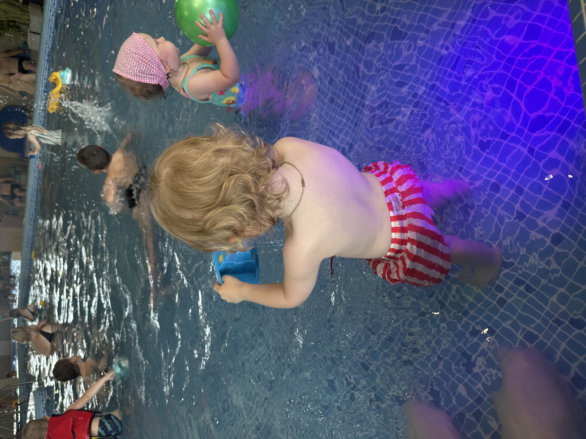 The photograph captures an indoor swimming pool scene, likely in Votkinsk, Russia, given the context. The pool features tiled walls and floor with water reflecting overhead lighting. Several individuals are present in the pool.
A young child with blonde, curly hair, wearing red and white striped swimming trunks, is positioned centrally in the frame, holding a blue toy container. Nearby, another younger child in a patterned swimsuit and a pink bandana holds a green ball. Other swimmers are visible in the background. The water is clear enough to see the pool’s tiled floor in places. The photograph seems to have been taken from an elevated position.