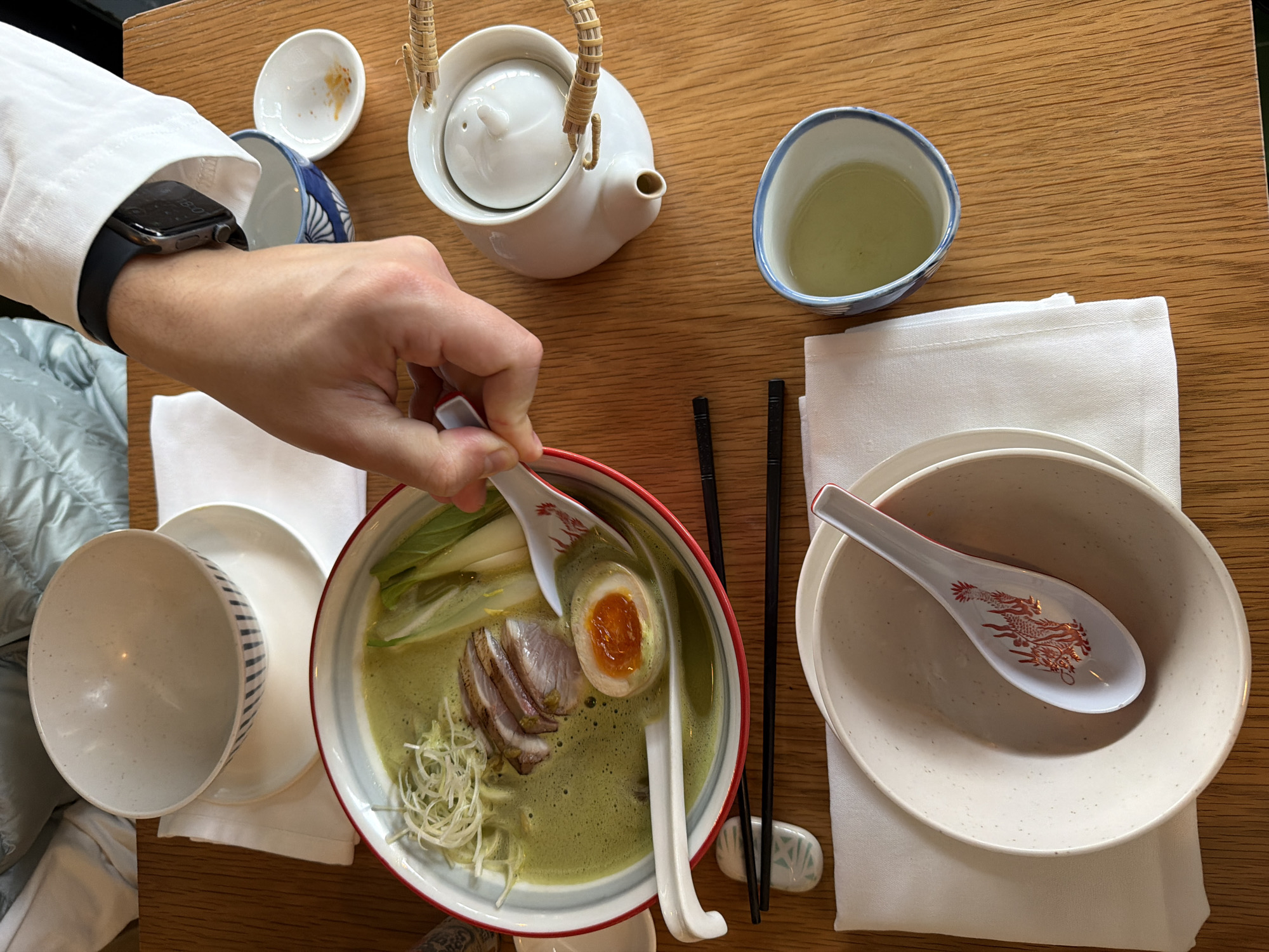 The image presents a top-down view of a meal setting on a light wooden table, likely in a restaurant in Tel-Aviv, Israel. A person's left arm, wearing a white long-sleeved shirt, is visible on the left side, with their hand holding a white ceramic spoon in a large bowl of ramen.
The ramen bowl, featuring a red rim, contains a vibrant green broth, several slices of meat (possibly pork belly), a soft-boiled egg with a rich orange yolk, green bok choy or similar vegetables, and shredded white scallions or leeks. The spoon held by the person also has a red border and a faint design.
To the right of the ramen, a pair of black chopsticks rests on a small, patterned ceramic chopstick holder. Further right, another white ceramic spoon, decorated with a prominent red dragon design, lies inside an empty white bowl which sits on a plain white plate and a folded white cloth napkin.
On the upper part of the table, a white ceramic teapot with a wicker handle is placed next to a small, empty white dish. A blue and white patterned teacup, filled with light green tea, is positioned in the upper right quadrant. On the left, a white bowl with blue striped exterior is inverted over a plain white plate, both resting on another white napkin.
The person's wrist wears a dark-colored Apple Watch, its screen displaying the time "18:01" (6:01 PM). The scene captures a moment of dining, highlighting a beautifully prepared bowl of ramen and accompanying tea.