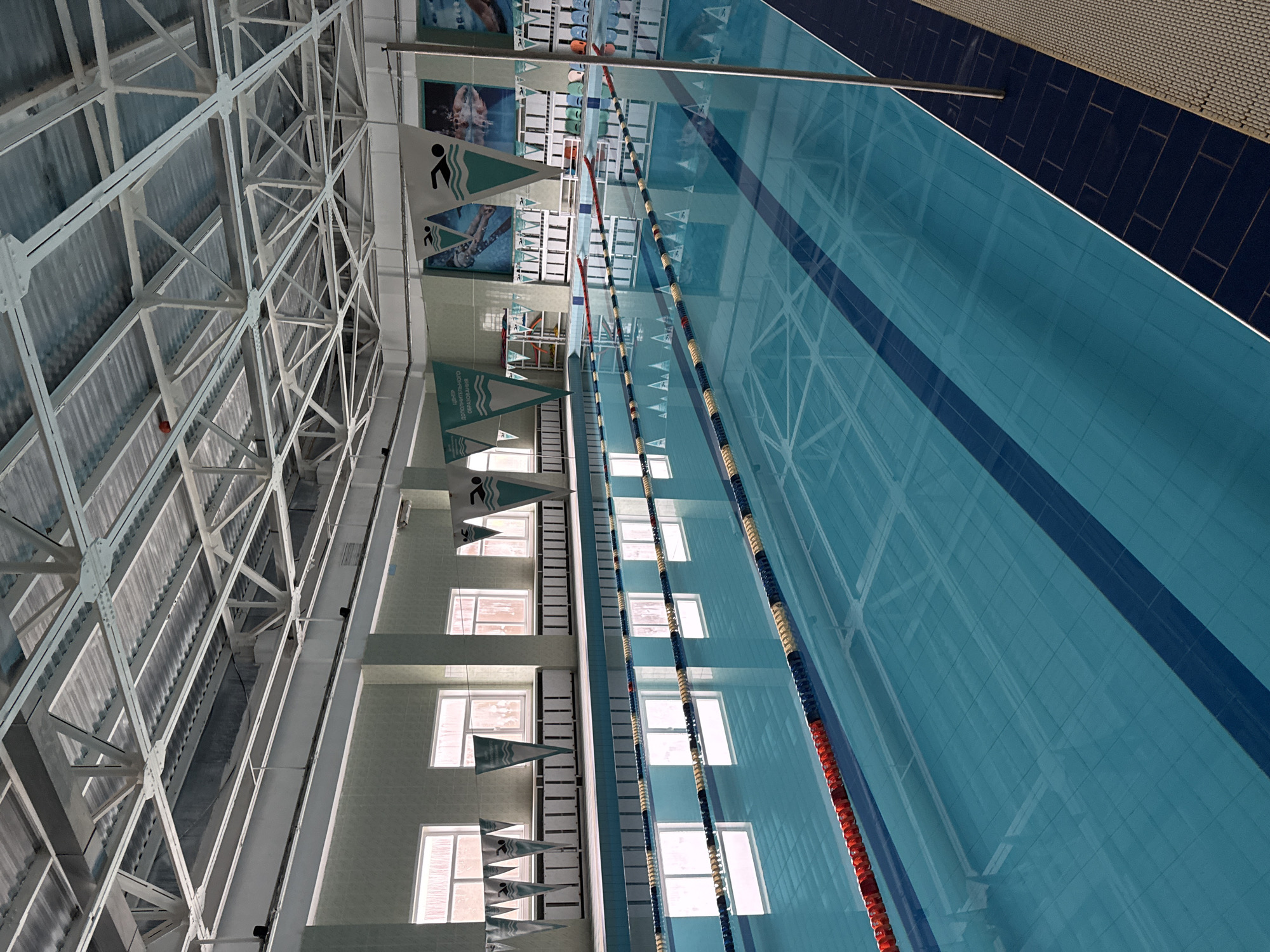 An indoor swimming pool in Markovsky, Russia, features multiple lanes marked by buoys and lane lines. The pool water is clear and blue, reflecting the ceiling structure and windows. The surrounding area includes tiled walls, benches, and decorative banners depicting swimmers. The architecture is modern, with a high, exposed metal ceiling structure and large windows. The lighting appears to be a mix of natural light from the windows and artificial overhead lighting. There are no people visible in the main area of the pool, but a single person is visible in the distance on the right side of the image.
