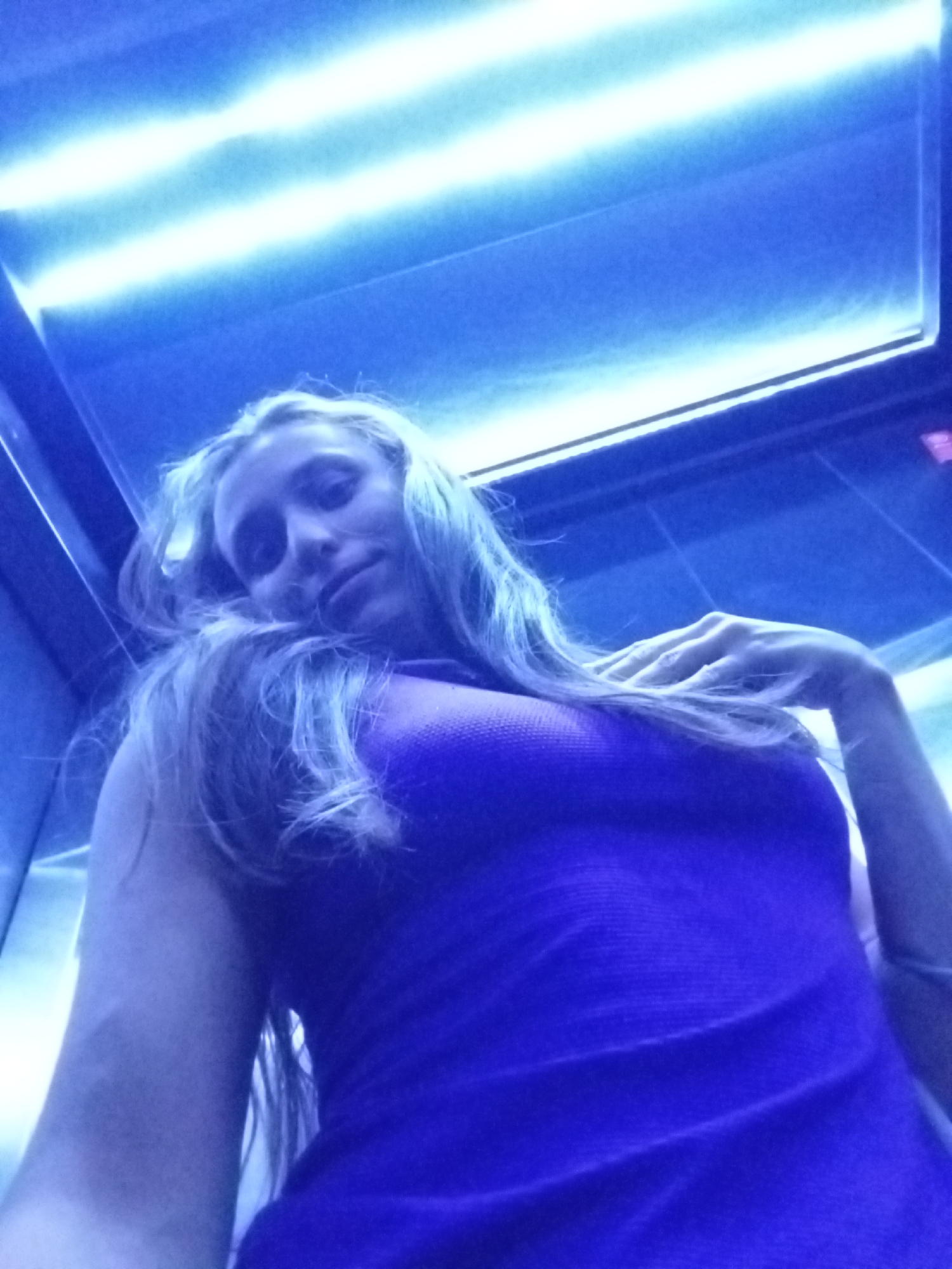 A woman with long, blonde hair is taking a selfie inside an elevator. She is wearing a purple tank top. The elevator's ceiling has fluorescent lights, casting a blue hue over the scene. The interior of the elevator appears to be stainless steel or a similar metallic material. The location in Buenos Aires, Argentina, is unknown.