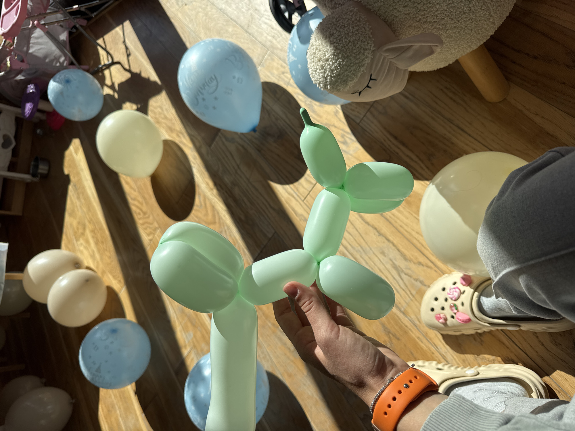 The image depicts a room with a wooden floor scattered with light blue and pale yellow balloons. A plush sheep toy is present in the upper right corner. In the center, a hand holds a green balloon animal shaped like a dog. A person wearing gray sweatpants and tan clogs adorned with charms is visible in the lower right corner, along with an orange smartwatch band. The scene likely represents a celebration, possibly a birthday. The location in Sofia, Bulgaria, cannot be determined from the visual information provided.