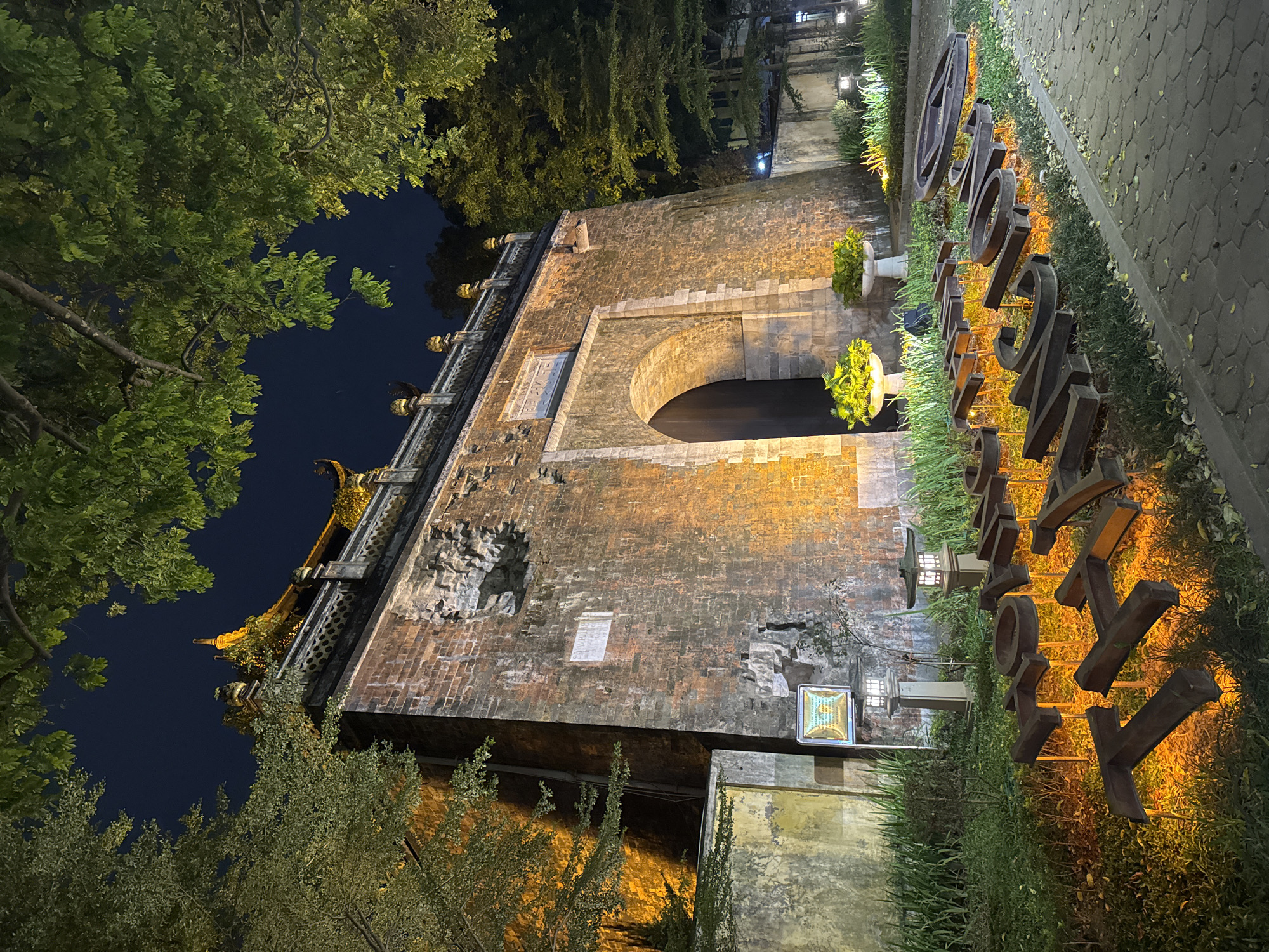 The image captures the imposing gate of the Imperial Citadel of Thăng Long in Hanoi, Vietnam, at night. The ancient brick structure is illuminated by warm, yellow lights, highlighting its arched entrance and weathered facade. Above the gate, a traditional tiled roof with intricate yellow detailing is visible against the dark night sky. Lush green trees frame the top of the image, their leaves illuminated by the ambient light. In the foreground, large, three-dimensional metal letters spelling out "HOÀNG THÀNH THĂNG LONG" (Imperial Citadel of Thăng Long) are lit from below, casting an orange glow onto the surrounding grass and pathway. The ground is paved with hexagonal stones. Two ornamental lanterns and potted plants add to the landscaped area in front of the citadel. The scene is serene and atmospheric, conveying a sense of history and grandeur.