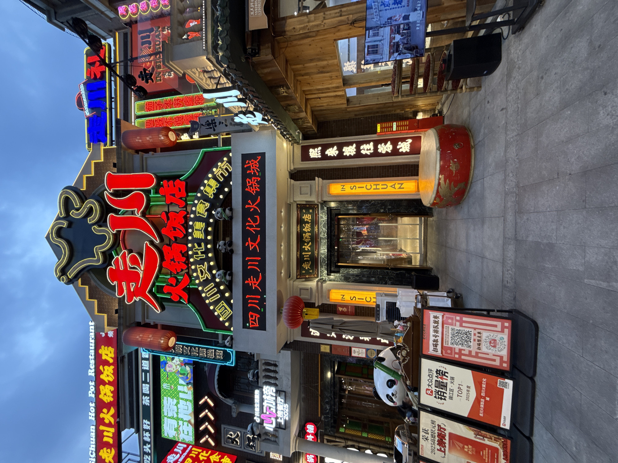 The image captures the illuminated facade of a multi-story Sichuan hot pot restaurant located in Jinjiang District, China. The central building features a prominent, ornate entrance adorned with numerous bright neon signs in red, green, blue, and yellow. The largest sign in the center displays the restaurant's name, "走川 火锅店" (Zǒuchuān Huǒguō Diàn), in large red Chinese characters, with an accompanying "SiChuan Hot Pot Restaurant" in English. A horizontal banner below the main sign reads "四川 走川 文化 火锅" (Sìchuān Zǒuchuān Wénhuà Huǒguō), translating to "Sichuan Zou Chuan Culture Hot Pot." Vertical light-up panels on either side of the entrance display "IN SICHUAN."
The architectural style is traditional Chinese, with decorative eaves, columns, and balconies. Several small panda figurines are placed on ledges and balconies across the facade. Two large red Chinese lanterns hang near the main glass entrance doors. To the left of the entrance, a large white panda sculpture with green bamboo accents is partially visible behind three A-frame display boards. These boards feature QR codes, promotional content, and mentions of "大众点评" (Dàzhòng Diǎnpíng), including a "销量榜 TOP1" (Sales Rank TOP1) for hot pot in "锦江区·火锅" (Jinjiang District Hot Pot) for 2023, and "2025高德优选榜" (Gaode Preferred List 2025).
To the right of the entrance, a large red ceremonial drum with gold dragon motifs rests on the grey tiled pavement. Beside it, a multi-tiered wooden rack holds an abundance of dried red chili peppers. Further right, a television screen on a stand displays images of what appears to be a traditional street scene or promotional video, with a black speaker positioned below it. The sky above is a muted twilight blue. Adjacent buildings, also featuring numerous brightly lit signs, are visible on both sides, indicating a bustling commercial or entertainment district. No people are clearly discernible in the foreground, suggesting a quiet moment on what is otherwise a vibrant street.
