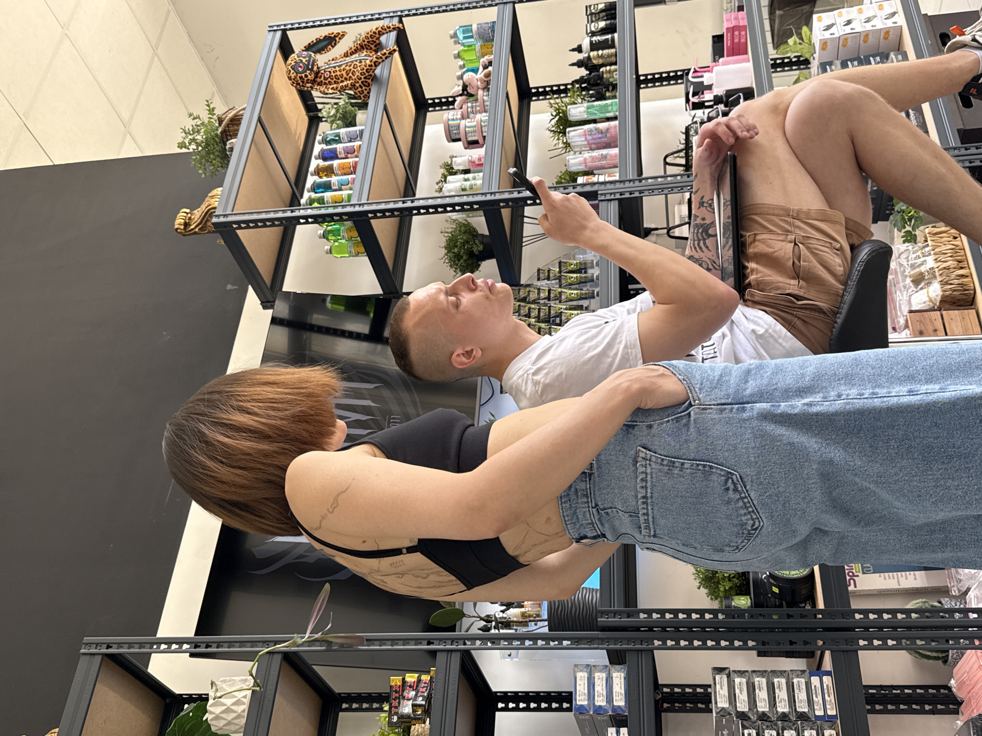 A woman with short brown hair wearing a black top and blue jeans stands facing left, seemingly embracing a man lying on his back in an elevated chair. The man, who has short, light-colored hair, wears a white shirt and tan shorts and holds a phone in his right hand. They are surrounded by metal shelving units filled with various products and decorative plants. The location is a store, presumably in Rostov-on-Don, Russia.
