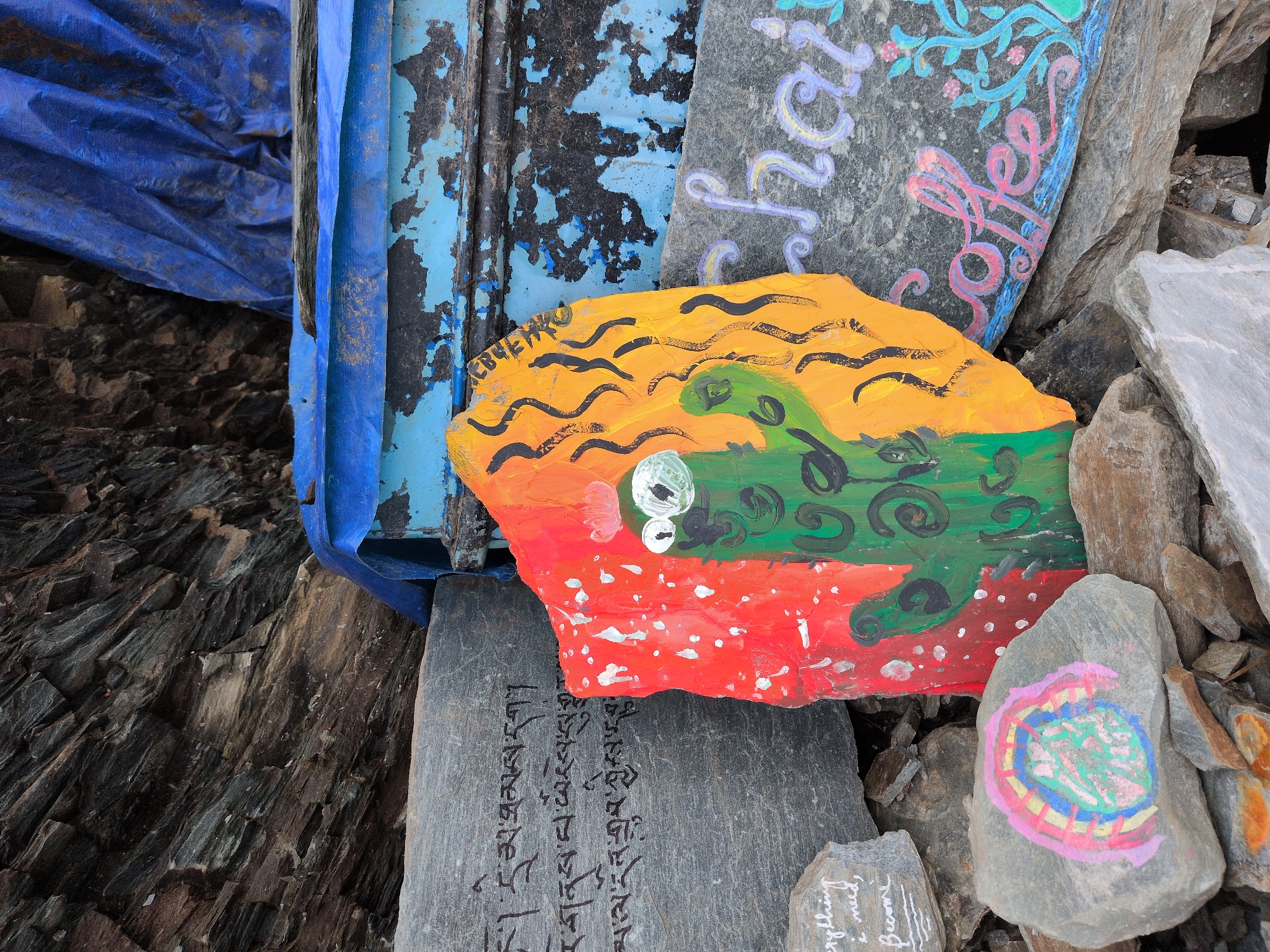 The image shows a close-up of several rocks with painted designs in Bhagsu, India. In the center, a flat, irregular-shaped rock is painted with a cartoonish green cactus wearing a red crown, set against a background of red on the left and yellow on the right. The cactus has large white eyes with black pupils. Black wavy lines are painted on the yellow section. To the left of the cactus rock, a larger, flatter grey rock has Tibetan or Buddhist script in black ink. Below this rock and to the left of the cactus rock, a smaller grey rock has the words "Everything I need," and "become" painted in white script. To the right of the cactus rock, another grey rock is painted with the word "chai" in purple script above the word "coffee" in pink script, with blue and green floral designs surrounding it. The rocks are situated amongst other natural rocks and rubble. In the background, a blue tarp is visible, along with a distressed blue and black structure. The overall impression is one of artistic expression in a natural, possibly rugged, outdoor setting in Bhagsu.