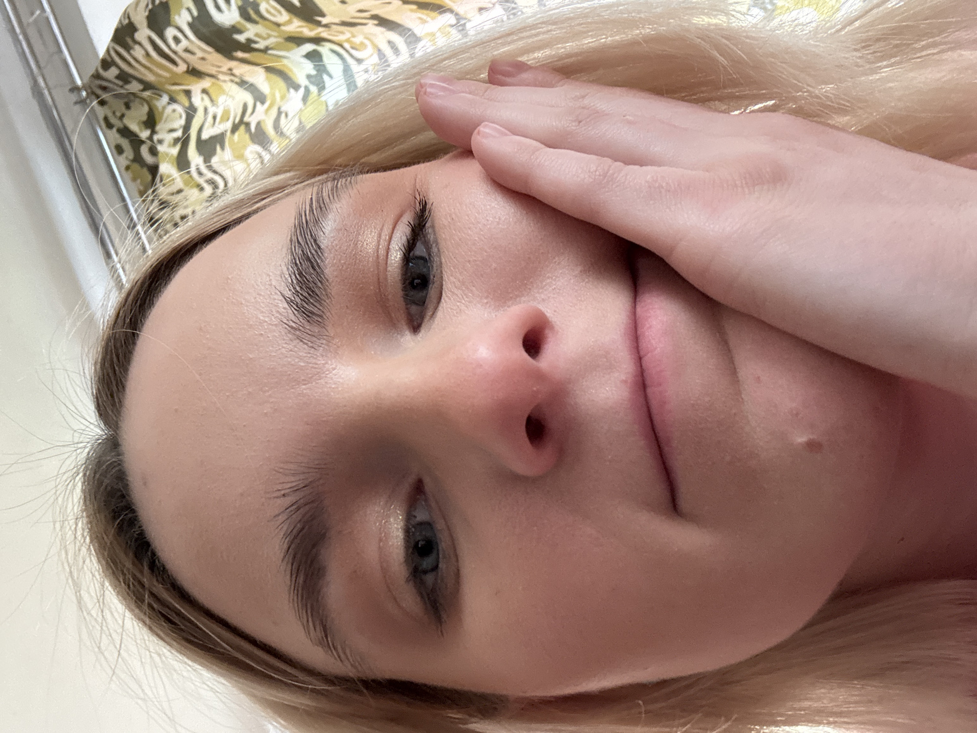 A close-up, angled selfie of a fair-skinned individual with blonde hair. The person is facing the camera with their right hand partially covering their face. The background shows a patterned surface. The location is Unknown, Unknown.