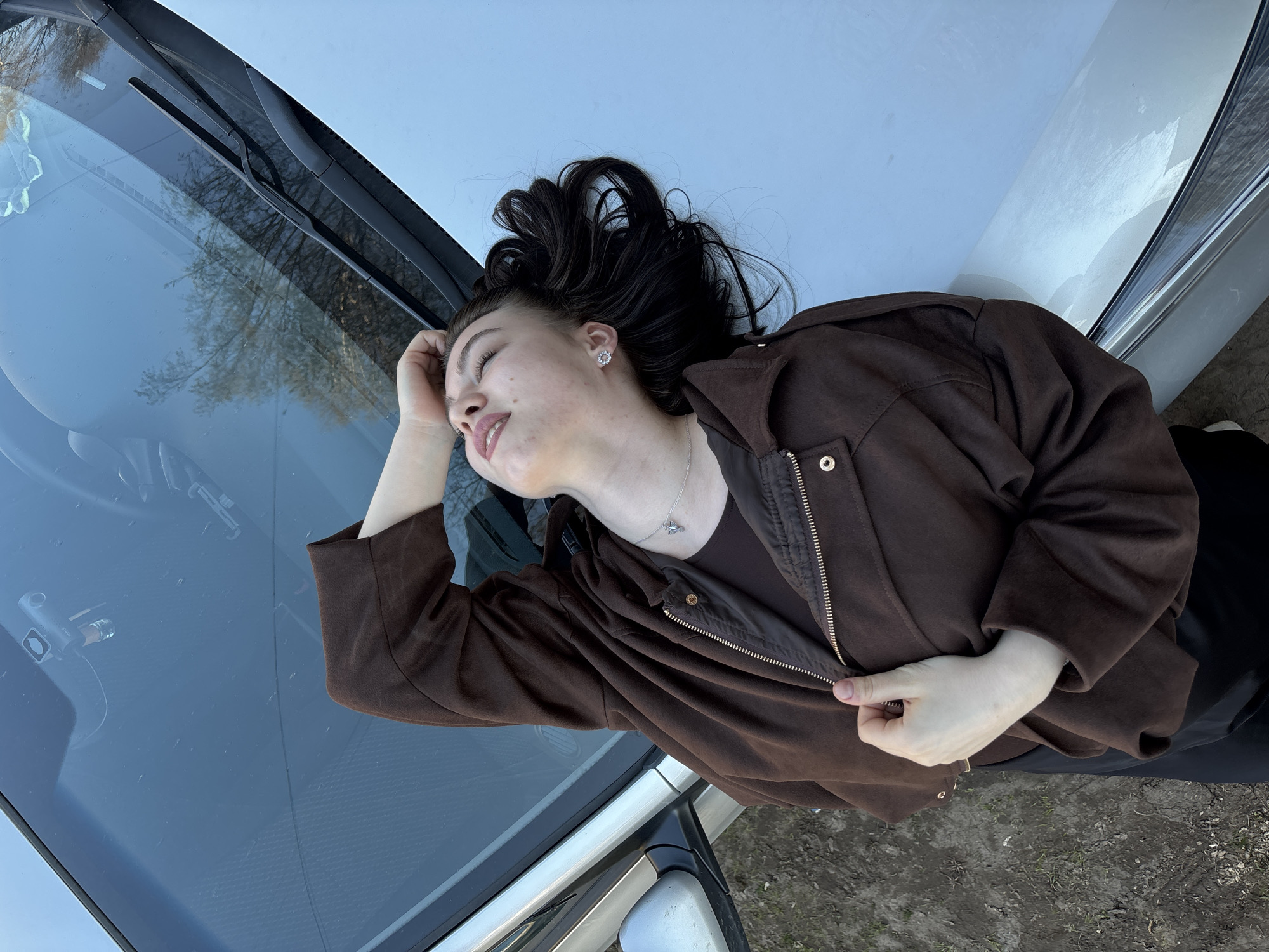 A young woman is lying on the hood of a light-colored car. She has dark hair and is wearing a brown jacket over a dark top, with black pants. Her left hand is resting on her forehead. The car's windshield and wiper blades are visible. The ground is partially visible, with some dry vegetation. The scene is outdoors, potentially in Perm, Russia, judging from the license plate visible in a crop and the presence of local flora.
