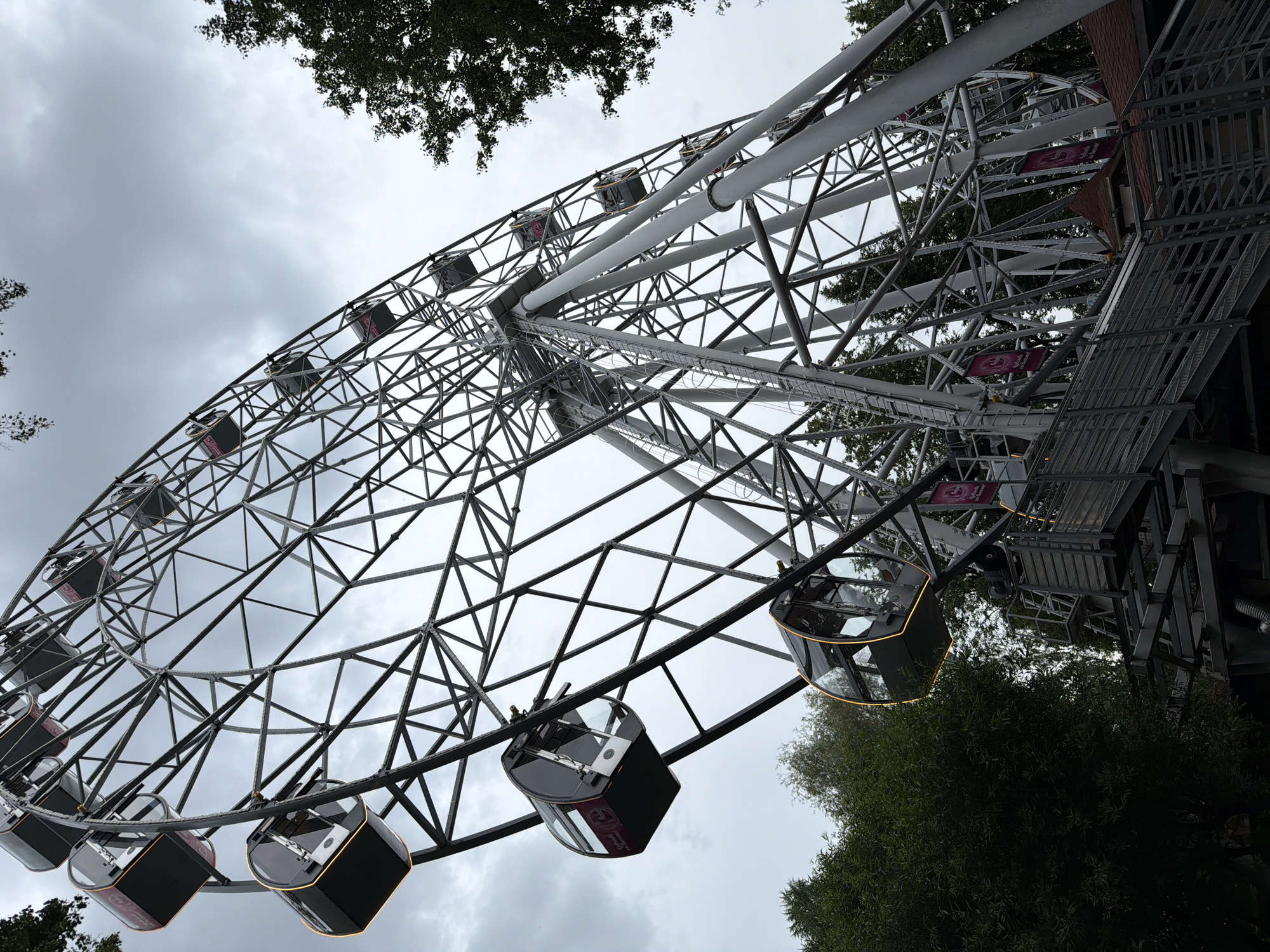 The image shows a Ferris wheel, a large amusement ride, located in the city of Perm, Russia. The Ferris wheel has a complex metal structure with multiple cabins suspended around its circumference. Some of the cabins are visible, appearing dark with illuminated windows and red accents. The ride is situated amidst trees and foliage, suggesting an outdoor park or recreational area. The sky is overcast with gray clouds, indicating a cloudy or possibly rainy day. The perspective is from below, looking up at the towering structure of the Ferris wheel. There are no visible people in the image. The scene captures a typical amusement park attraction under a diffused light condition.