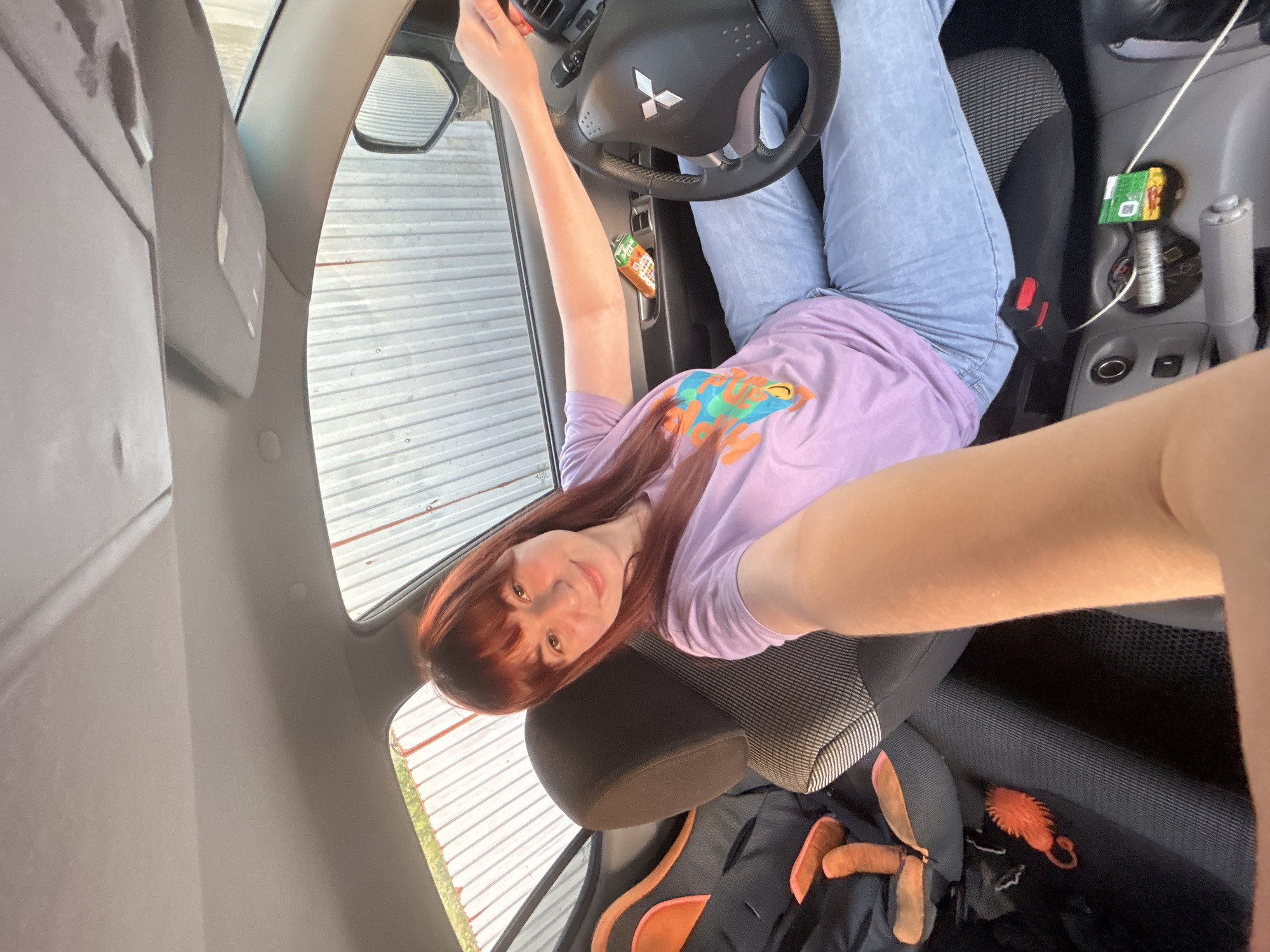 A young woman is taking a selfie inside a car. She has long, reddish-brown hair and is wearing a light purple t-shirt and light blue jeans. The car is a Mitsubishi, as indicated by the logo on the steering wheel. A juice box is visible in the door storage. A backpack with orange accents is on the passenger seat. There is no indication this image was taken in the city of Фоки, Russia.