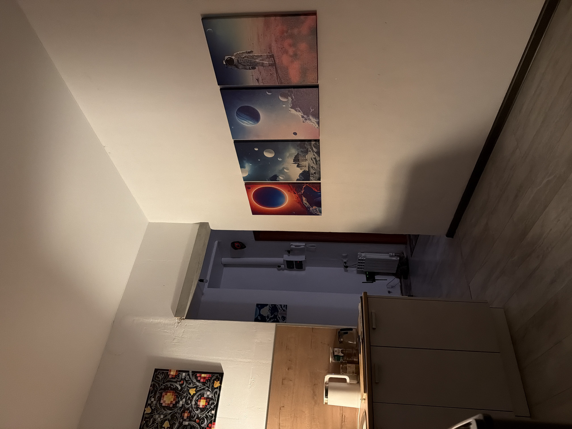 The image shows an interior scene, likely a hallway or kitchen area, with several pieces of art displayed on the wall. The artwork consists of four rectangular panels arranged horizontally, depicting space-themed imagery with planets, nebulae, and an astronaut.
On the left side of the image, a kitchen counter is visible with a white electric kettle, jars, and what appears to be a coffee maker. Above the counter, a framed piece of art with a geometric pattern is hung on the wall. The walls are painted white, and a doorway leads into a darker area.
The flooring is light-colored laminate. The lighting in the scene is dim, casting shadows and creating a warm ambiance. The overall impression is that of a lived-in domestic space. The specific location within Naberezhnye Chelny, Russia, cannot be determined from the visual information provided in the image.