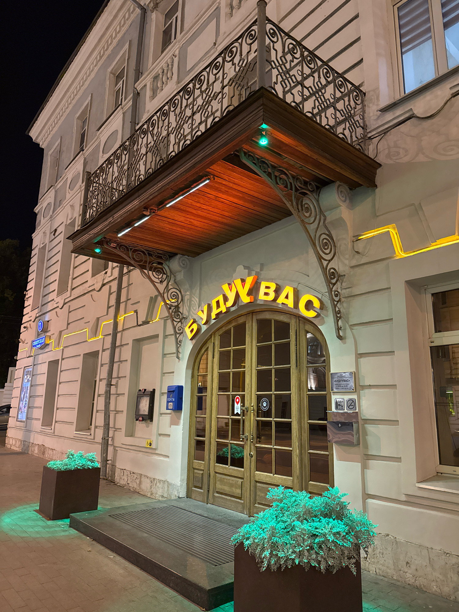 The image shows the entrance of a building at night, located in Tver, Russia. The building has a light-colored facade with ornate details and a wrought-iron balcony on the second floor. Above the entrance, a wooden overhang is illuminated by warm lights. Large, arched double doors made of wood and glass lead into the building. A yellow neon sign above the doors spells out "БУДУ ВАС" in Cyrillic letters.
To the left of the entrance, there is a planter with green foliage, illuminated by green lights from below. To the right of the entrance, there is a mailbox and a sign indicating the establishment's name and operating hours. A sidewalk runs in front of the building, and a street is visible in the background, illuminated by streetlights. The overall atmosphere is dimly lit and nocturnal, suggesting the establishment is a restaurant or bar. The building's architecture appears to be historic.