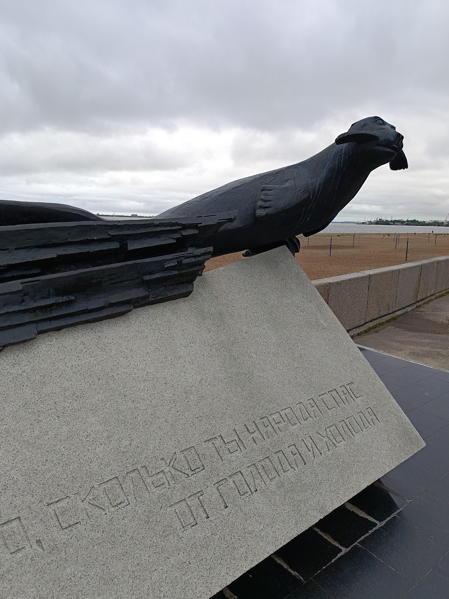 This is a medium-angle, eye-level shot of a monument in Arkhangelsk, Russia, situated on a waterfront. The monument features a large, dark bronze sculpture of a seal with a fish in its mouth, depicted in a dynamic pose as if emerging from or resting on a layered, abstract form. The sculpture is mounted on a large, light-colored granite pedestal with engraved text in Russian. The text, partially visible, reads "СКОЛЬКО ТЫ НАРОДА СПАС ОТ ГОЛОДА И ХОЛОДА" which translates to "How much of the people did you save from hunger and cold."
The background shows a sandy beach, a concrete embankment wall, and a body of water with industrial structures and a hazy skyline in the distance. The sky is overcast with heavy, gray clouds. The ground in front of the pedestal is paved with dark, reflective tiles. There are no people clearly visible in the immediate foreground or midground, but a distant figure can be seen on the beach. The overall mood is somber and commemorative, enhanced by the overcast weather and the subject matter of the monument.