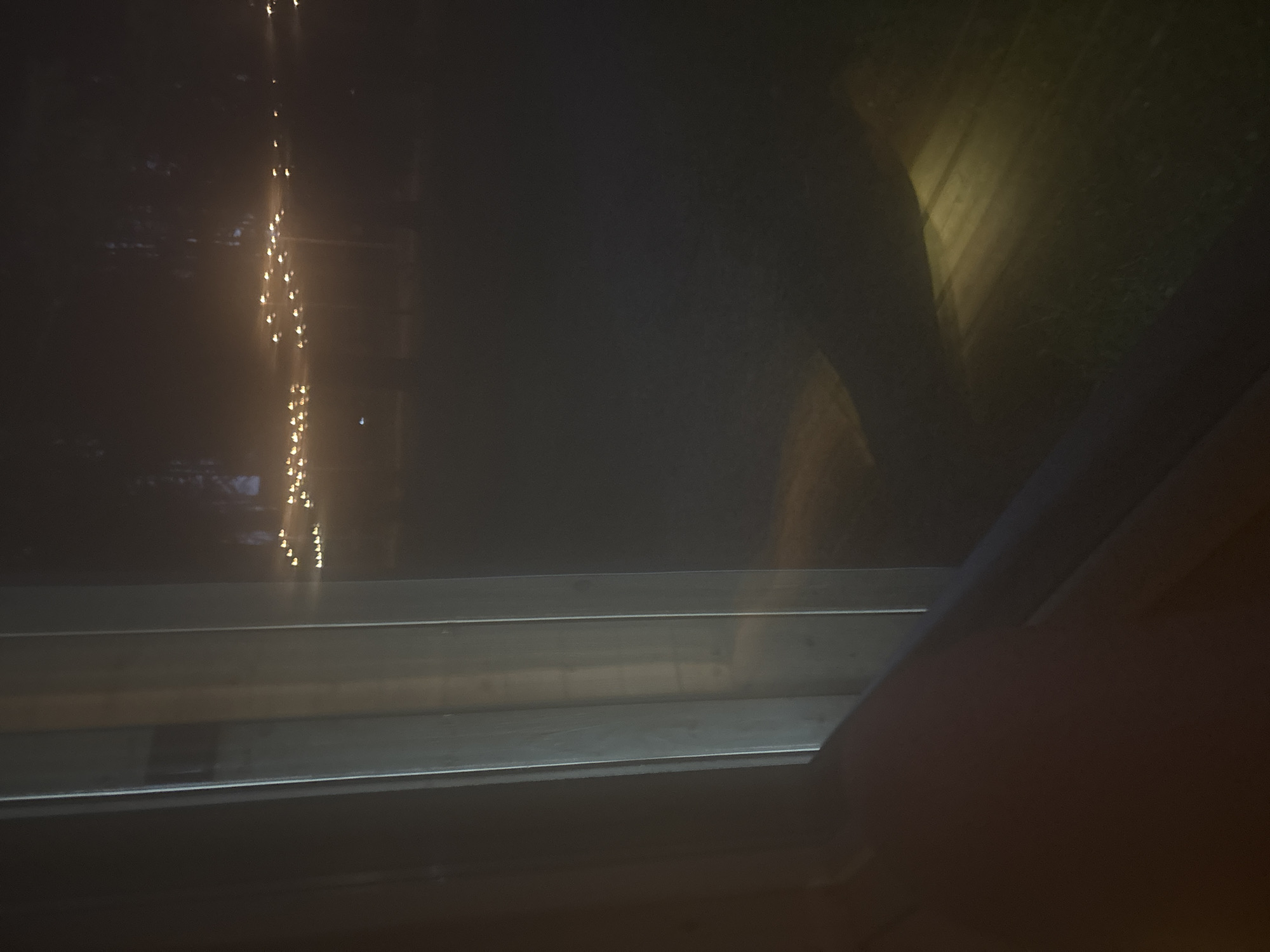 The image displays a nocturnal outdoor scene viewed from an interior perspective in the city of Мишкино, Russia. On the left, two prominent vertical, light-toned window mullions exhibit subtle reflections. A dark, curved form partially obscures the lower left frame, consistent with a human arm or shoulder. The exterior is dark, featuring a horizontal line of numerous small, amber-colored lights across the mid-ground, appearing slightly diffused due to distance or atmospheric conditions. Faint, dark vertical silhouettes, consistent with trees, are discernible in the background above these lights. Two distinct, elongated, yellowish light beams are projected diagonally onto the dark ground in the lower right and center, illuminating a textured surface indicative of earth or sparse vegetation. No explicit human actions or interactions are discernible beyond the potential partial presence of a viewer's limb.