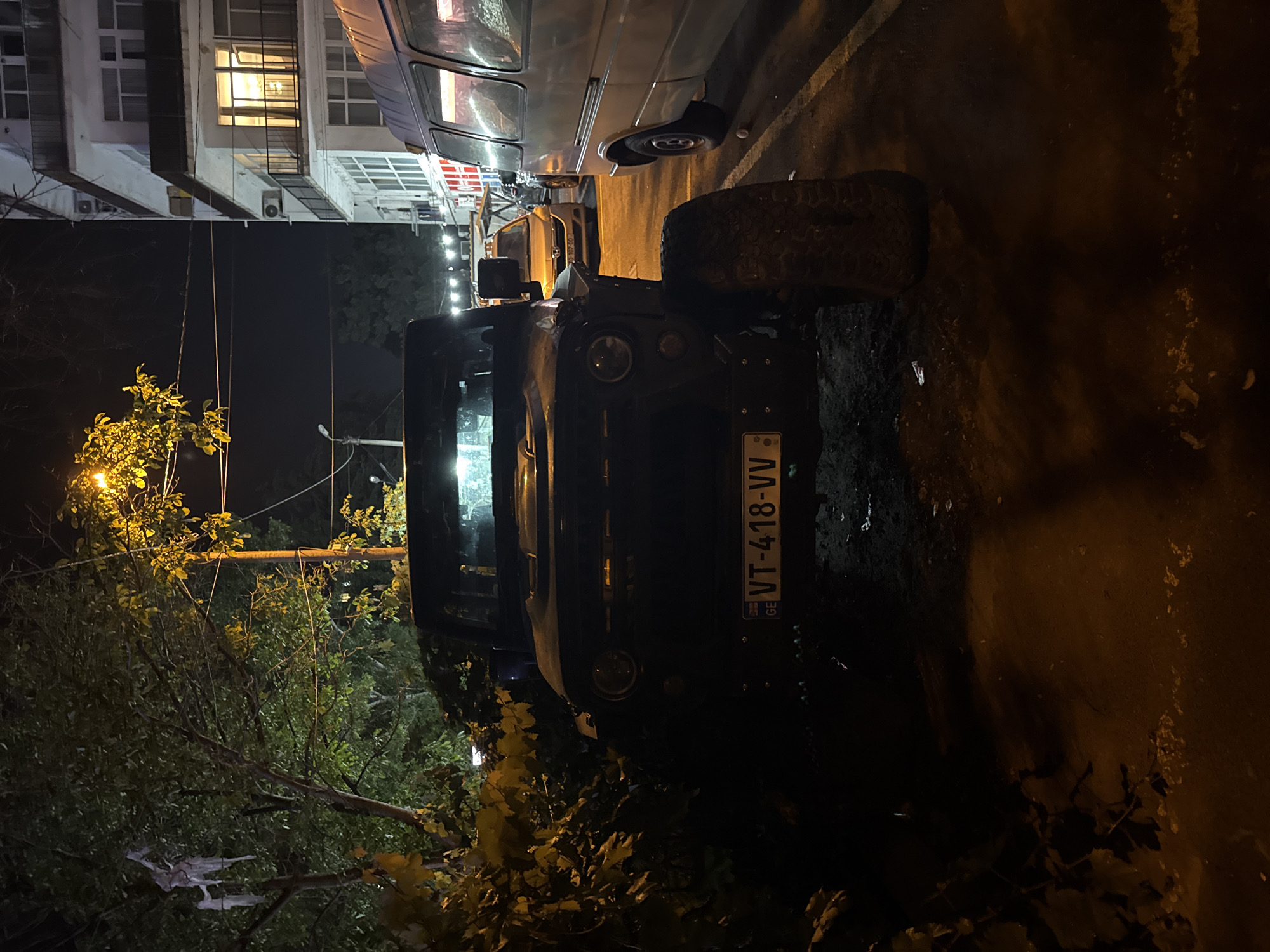 At night, a dark-colored off-road vehicle with a license plate reading "GE VT-418-VV" is parked on a dirt and gravel surface. The vehicle is partially obscured by dense foliage on the left side of the frame. To the right of the off-road vehicle is a light-colored van. In the background, a multi-story building with balconies and illuminated windows is visible. The scene is lit by streetlights casting a warm, yellow glow. The location is the city of Tbilisi, Georgia.
