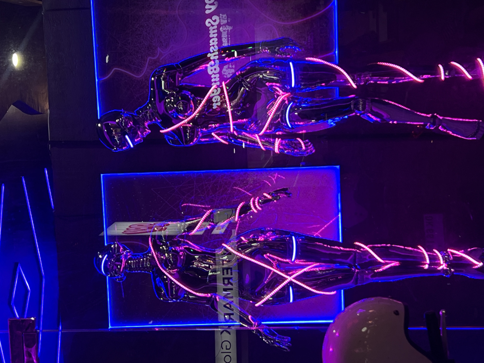 Two chrome mannequins, illuminated with purple and blue neon lights, are displayed in North Kuta, Indonesia. The mannequins are posed as if floating horizontally. Pink LED strips are wrapped around them in an intersecting pattern. Text is visible behind the mannequins, inverted and partially obscured. A white, dome-shaped object appears in the lower right corner.