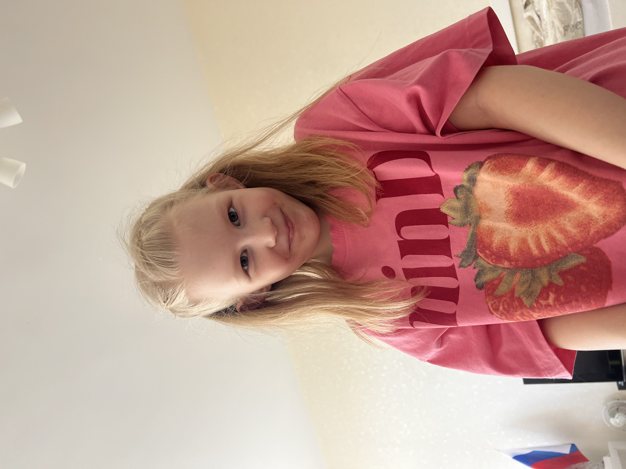 A girl with blonde hair is wearing a pink t-shirt with a strawberry print. The photograph appears to be taken indoors, possibly in a residential setting. The girl is smiling slightly. A wall and a ceiling are visible as the background. The location of the photograph is not evident from the image. There is no specific information to indicate this photograph was taken in Yekaterinburg, Russia.