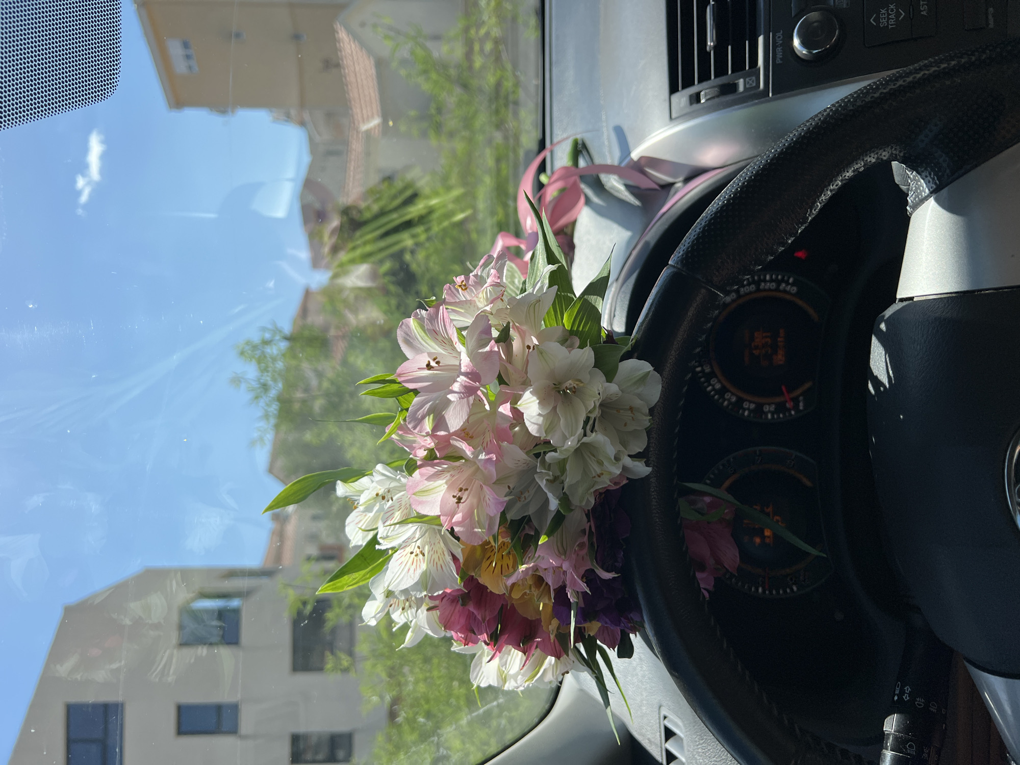 The image shows a bouquet of flowers positioned in front of the steering wheel and dashboard of a car. The flowers are a mix of colors including white, pink, purple, and yellow. A pink ribbon is visible amongst the flowers. The dashboard and steering wheel are black and silver. The windshield reflects the outside environment, showing a building with windows, trees, and a blue sky with clouds. The location in the city of Budva, Montenegro is not discernible from the image.