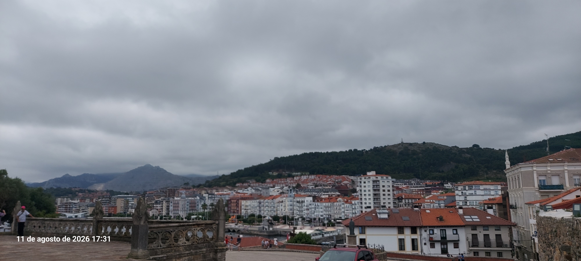 The image captures a panoramic view of the city of Castro-Urdiales, Spain, under a heavily overcast sky. In the foreground, a decorative stone railing with intricate carvings runs across the bottom of the frame. To the left, a person wearing a pink cap and light-colored clothing stands near the railing, facing away from the camera. Further left, another person in dark clothing is partially visible.
The middle ground displays a dense urban landscape with numerous buildings characterized by red-tiled roofs and a mix of white, grey, and muted-colored facades. Several taller buildings with white exteriors and numerous windows are interspersed among the lower structures. A body of water, likely a harbor or bay, is visible in the lower-middle section of the image, with boats docked and some activity on the waterfront.
In the background, rolling green hills rise, densely covered with trees. On the crest of one of the hills, a tall, slender structure, possibly a radio tower or monument, is discernible. The overall atmosphere is subdued due to the cloudy weather, which obscures any direct sunlight and creates a diffused light across the scene. A timestamp in the bottom left corner indicates "11 de agosto de 2026 17:31."