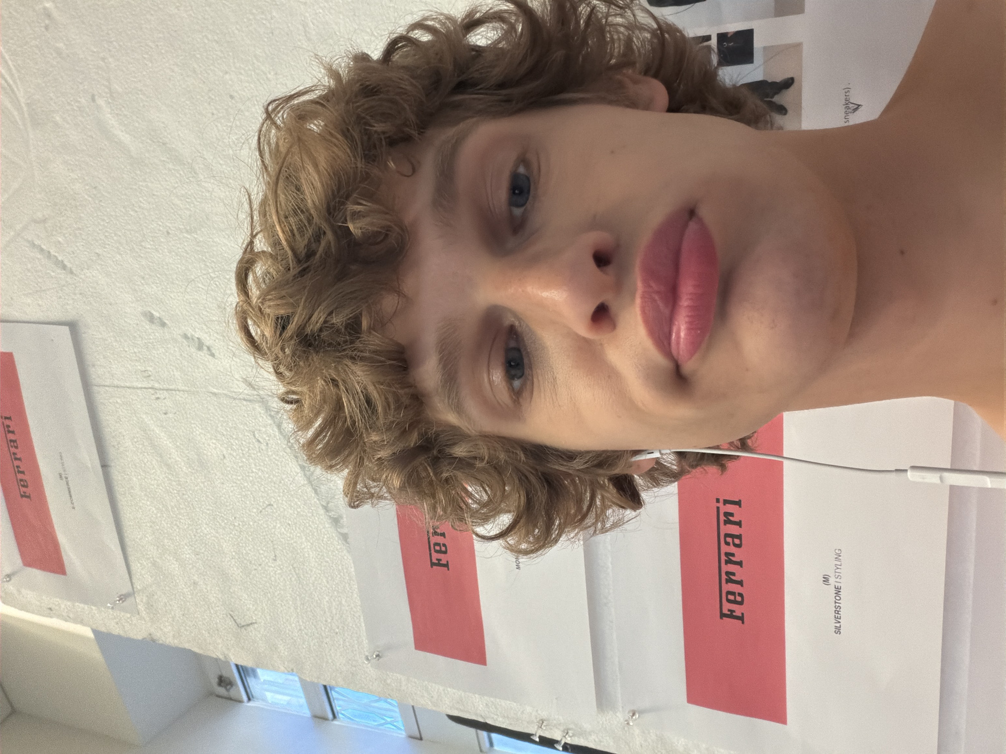 The photograph shows a person with curly blonde hair, fair skin, and blue eyes looking directly at the camera. The person's lips are pursed. The background is a white wall with posters or prints attached. The posters have a red stripe and the word "Ferrari" printed on them. The text "SILVERSTONE | STYLING" is visible on one poster. There is a window at the bottom edge of the frame. An earbud wire is visible. There is no information in the picture to confirm that the image was taken in Milan, Italy.