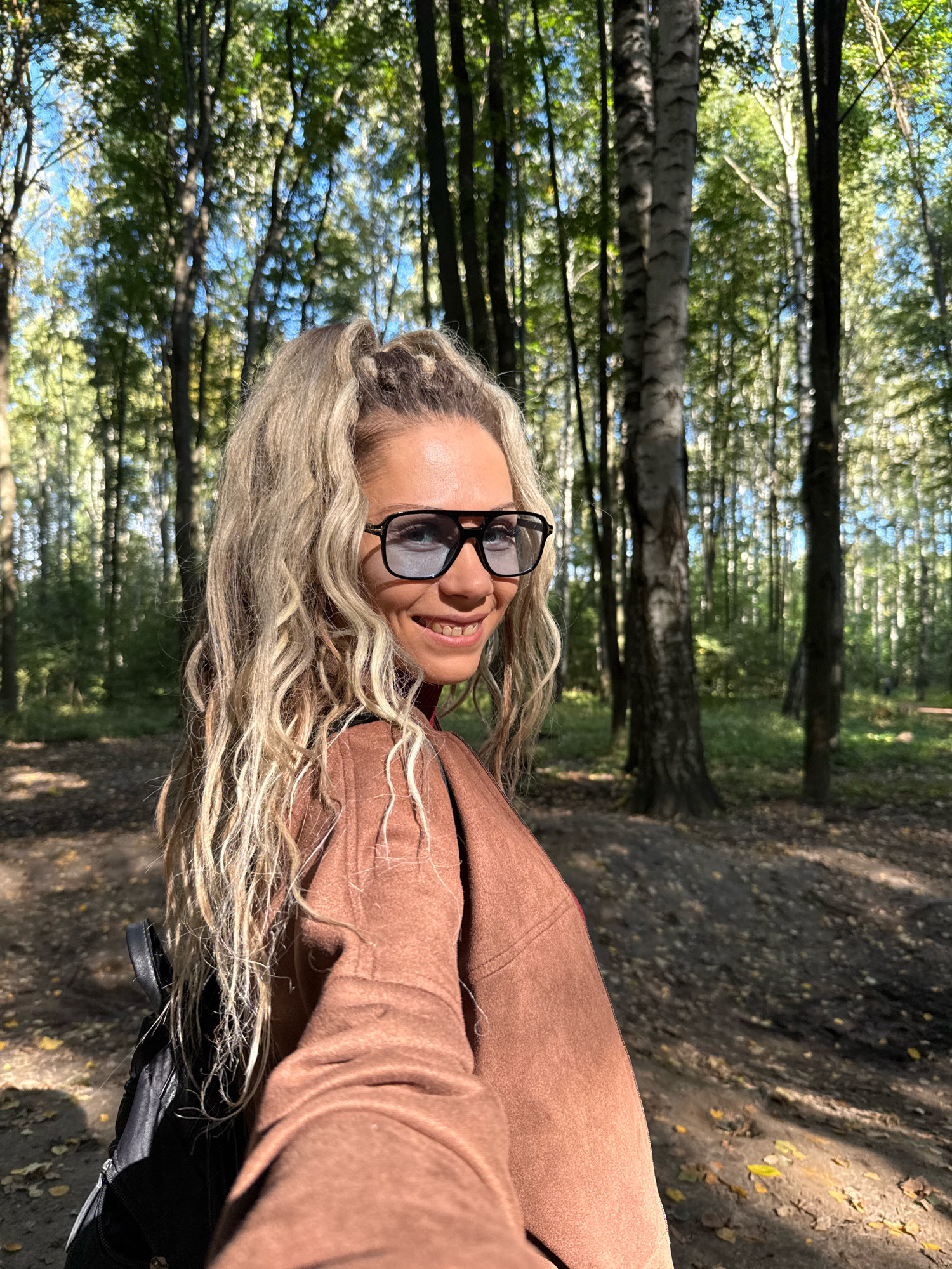 A medium shot shows a woman with long, wavy blonde hair pulled into a high ponytail. She is wearing dark-rimmed eyeglasses and a brown coat. The woman is outdoors in a forest, with trees and dappled sunlight visible in the background. The ground is covered in fallen leaves, suggesting it is autumn. She is looking towards the camera and smiling. The location is the city of Rusanovo, Russia.