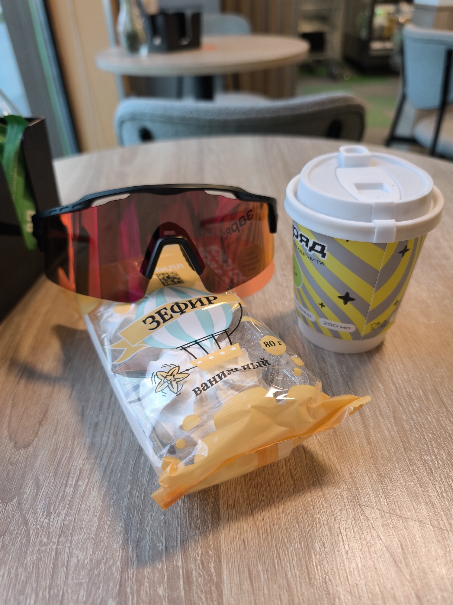 A coffee cup, a package of marshmallow candy, and a pair of sunglasses are on a table in a cafe. The coffee cup is white with a yellow band around it and black stars. The package of marshmallow candy is clear and says "Zefir" in Russian. The sunglasses are black with red lenses. The table is wooden. The city is Moscow, Russia based on the cup.