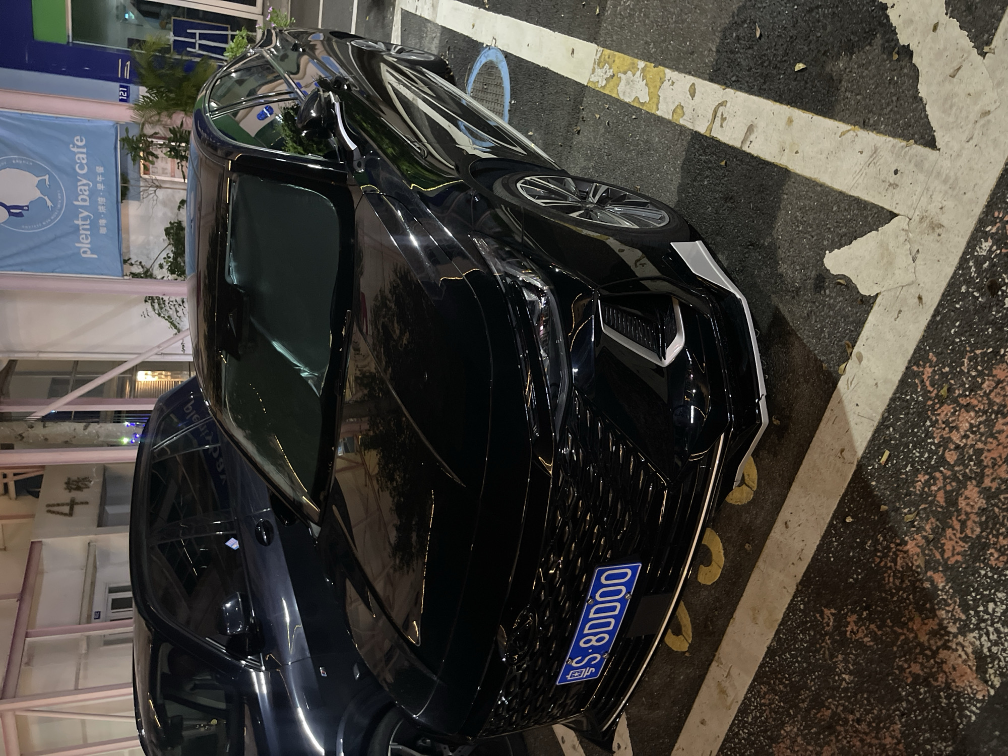 This media file shows a black car parked on a street at night. The car is a prominent object in the foreground, reflecting the surrounding lights. To the left of the car, a blue banner with white text announces "plenty bay cafe" and mentions "coffee, baking, brunch" in Chinese characters. Next to the banner, a building with the number "121" is visible. Further left, another building shows Chinese characters that translate to "Building 4". Yellow and white painted lines on the asphalt suggest a parking area. The overall scene is a street in Panyu District, China, at night. No people are visible, and the focus is on the car and the immediate surroundings.