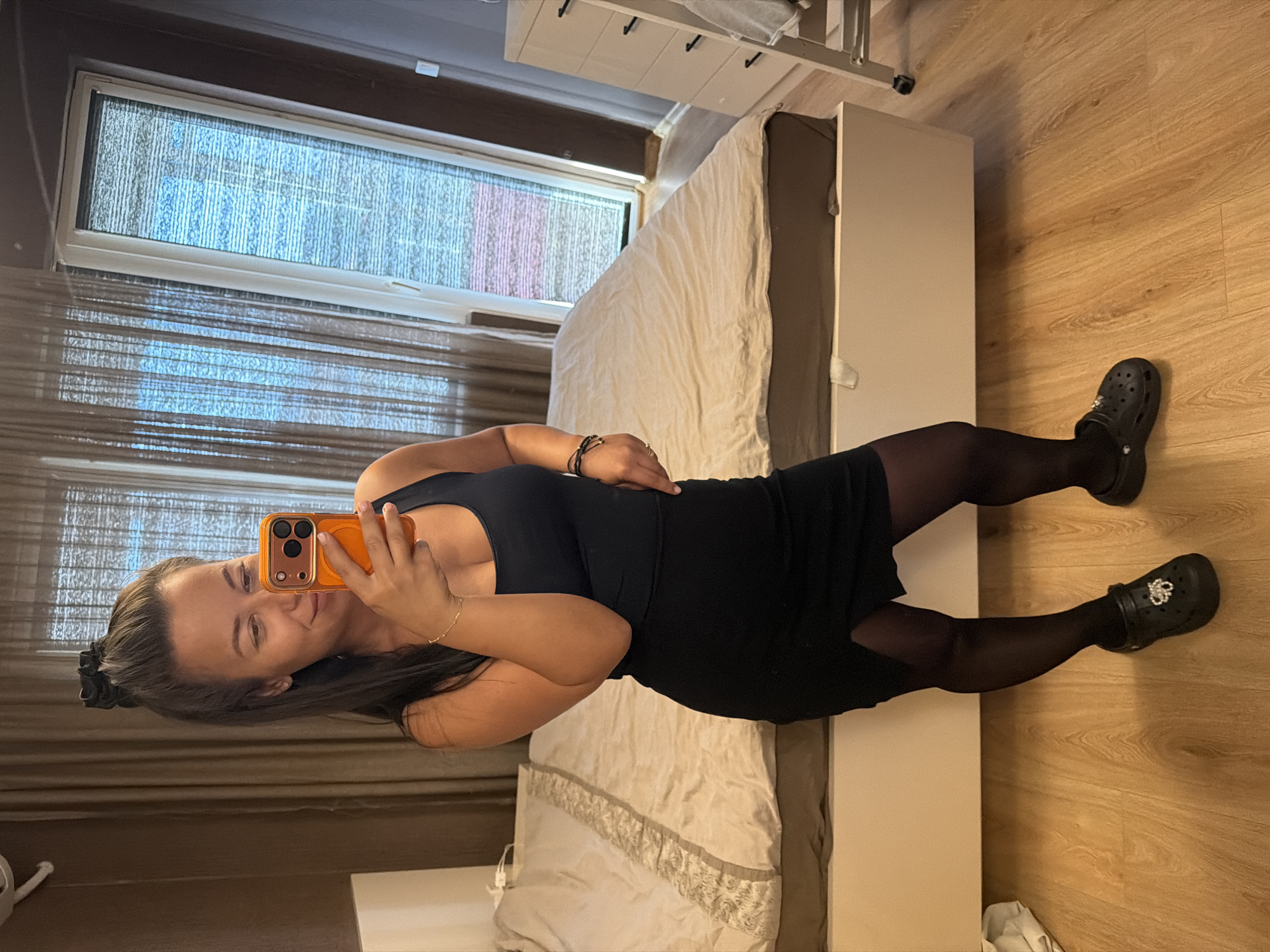 The image captures a full-body selfie taken by a woman indoors. She is standing in what appears to be a bedroom, with a bed visible behind her. The room has light brown curtains and a white dresser with black drawer pulls to the right. The floor is made of light-colored wood. The woman is wearing a black tank top, a black skirt with a slit, black tights, and black Crocs. She is holding an orange smartphone in her right hand, using it to take the picture. Her hair is pulled back in a high ponytail with a black scrunchie. She is looking at her reflection in the phone's screen and smiling. The image does not provide any specific visual cues to indicate a location in the city of Burgas, Bulgaria.
