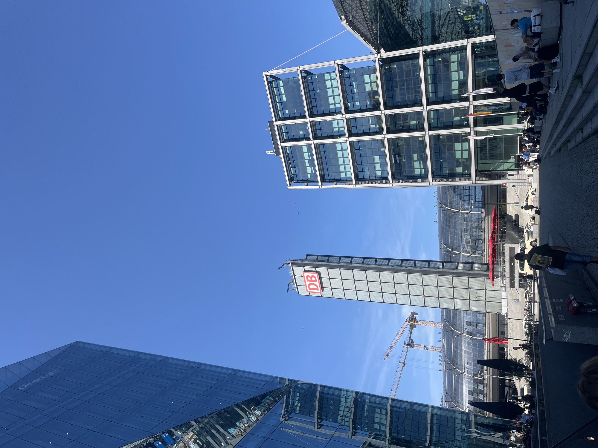 This image depicts a scene at Berlin Hauptbahnhof, Germany's largest and busiest railway station. The foreground shows a pathway with people walking and sitting, suggesting a public space. To the left, a modern glass-fronted building dominates the frame, reflecting the blue sky. In the center, a tall, slender tower with the prominent red "DB" logo stands against the clear sky. In the background, the glass roof of the train station is visible, along with construction cranes, indicating ongoing development or maintenance. Another contemporary building with a grid-like facade is situated to the right of the "DB" tower. The overall environment appears to be an urban setting with a focus on modern architecture and transportation infrastructure.