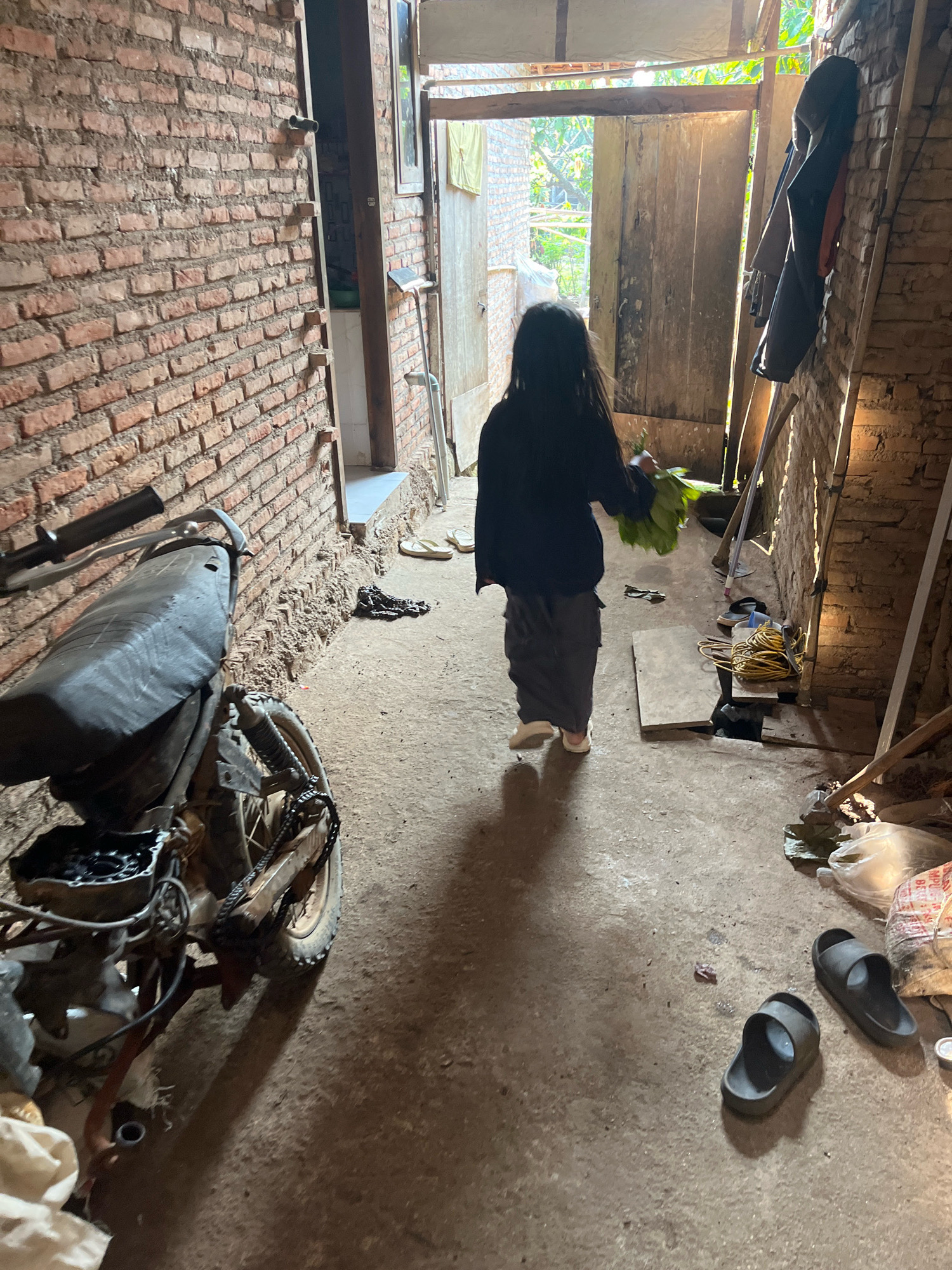 An outdoor scene in Kebumen, Indonesia, features a young child with long, dark hair walking away from the camera towards an open doorway. The child is wearing a dark top and dark pants, carrying a bunch of green leaves in their right hand. The environment appears to be a yard or alleyway with unfinished brick walls on the left and right. A motorcycle is parked on the left side of the frame. The ground is a mixture of concrete and dirt. Various objects are scattered around, including a pair of sandals, a coiled rope, and a plastic bag. The doorway leads to an area with natural light, suggesting an exit or a more open space beyond.