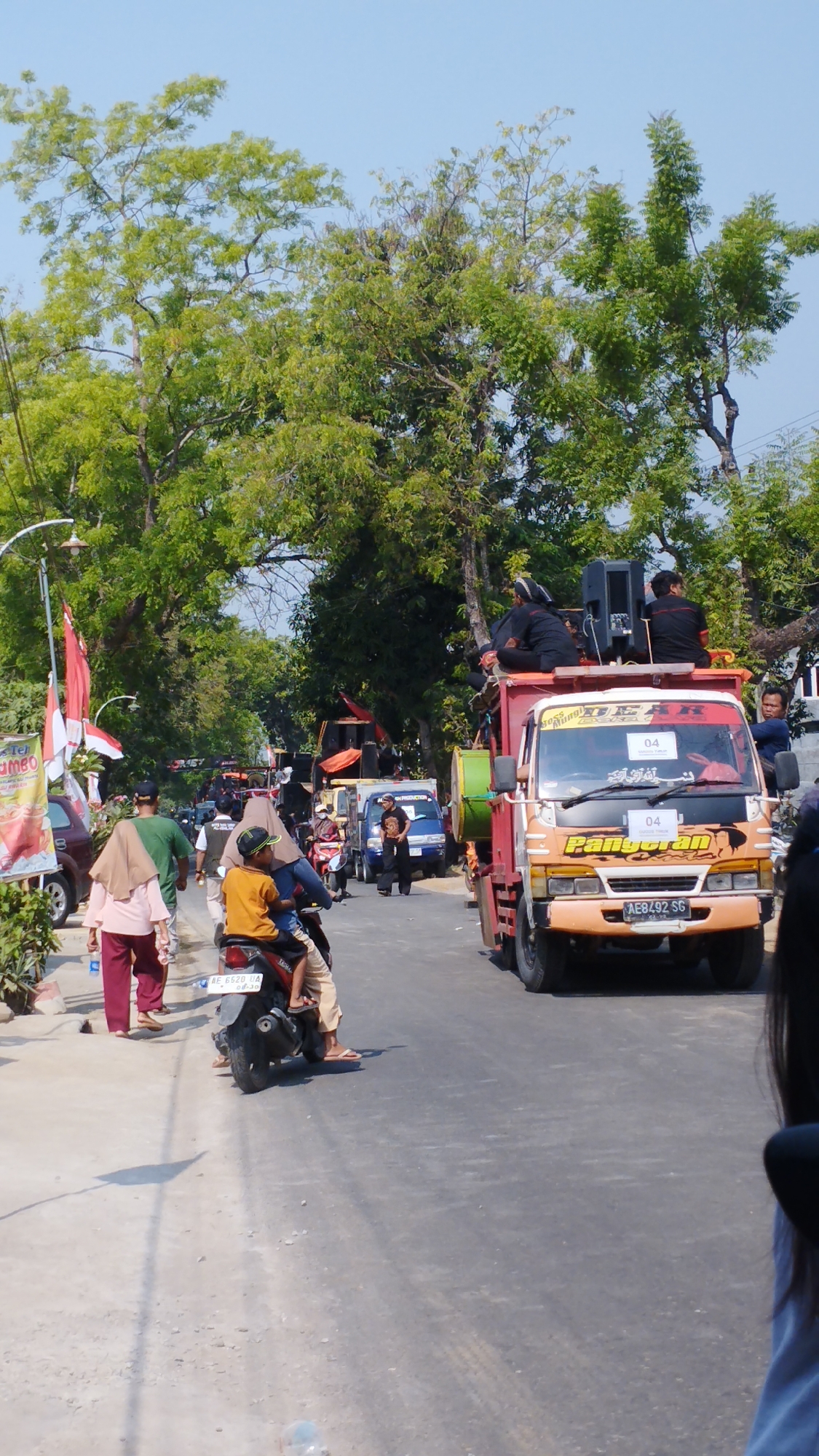 The image depicts a street scene in Bancar, Indonesia, with a procession or parade in progress. A flatbed truck, brightly colored in orange and red, is prominent in the foreground, carrying several individuals dressed in black attire and large speakers on its back. The truck has the number "04" displayed on a white sign. Beside the truck, a blue pickup truck also appears to be part of the procession.
On the left side of the street, pedestrians are walking, including a woman in a pink headscarf and pink pants, and a man in a green shirt. A young person in an orange shirt and a cap rides on the back of a motorcycle that is moving along the road. Indonesian flags, red and white, are visible along the side of the road, attached to poles and a white archway.
The environment consists of a paved road lined with trees providing ample shade. The sky is clear and blue. The overall impression is a lively event taking place on a sunny day in a semi-urban or town setting in Indonesia.