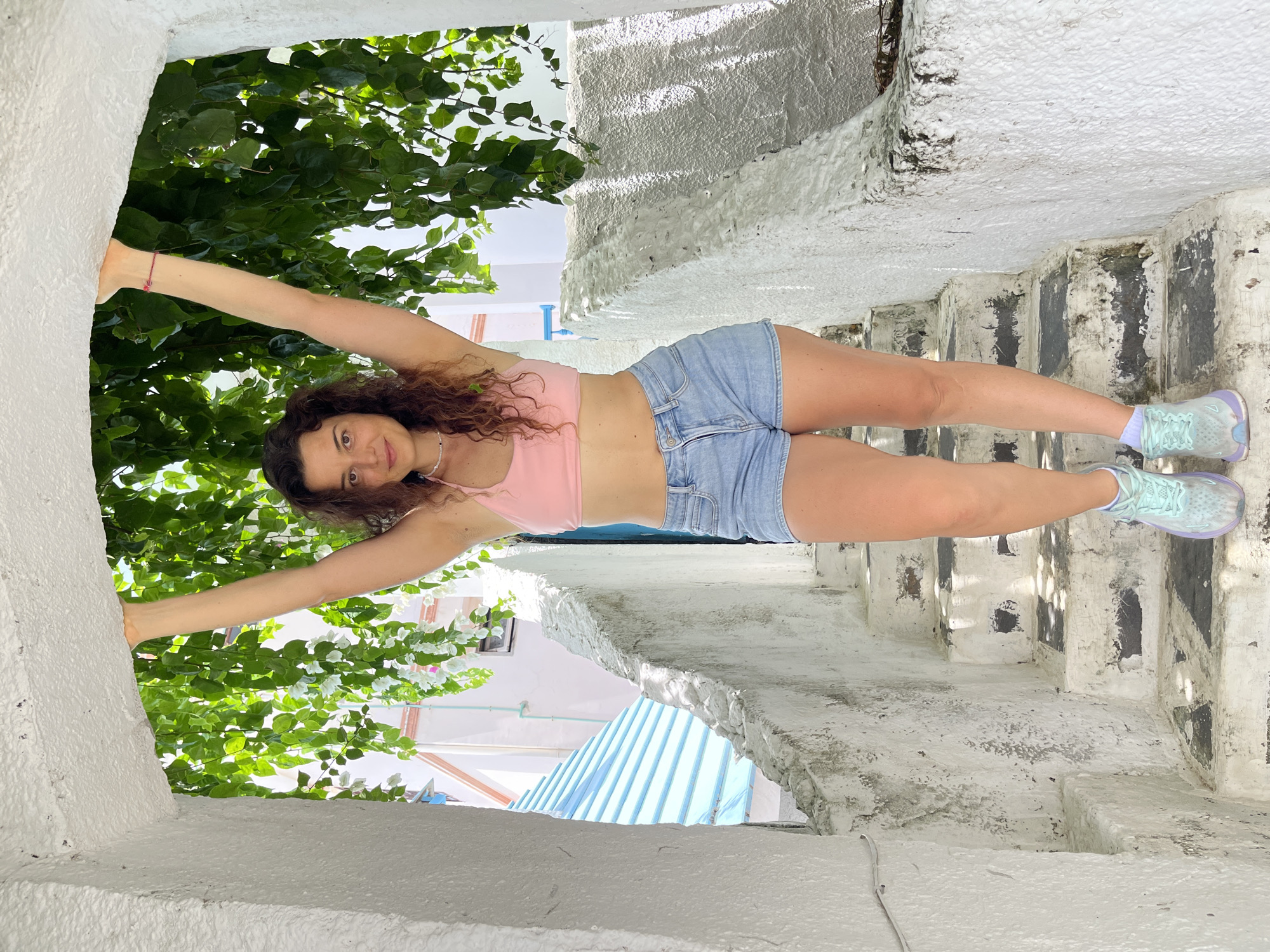 A woman stands on a set of outdoor stairs, holding onto an archway overhead. She has long, curly brown hair and is wearing a pink halter-neck crop top, denim shorts, and light blue sneakers. The stairs are made of concrete and painted white, with some patches of wear and tear. Lush green foliage hangs from above, partially obscuring the background. In the distance, a building with a blue roof is visible. The scene appears to be a narrow pathway or alleyway, possibly in a residential area. The lighting suggests it is daytime with sunlight casting shadows on the stairs.