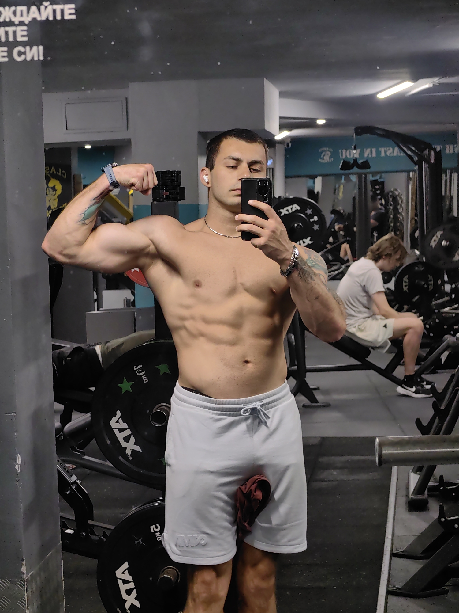 A young man in athletic wear poses for a mirror selfie in a gym. He has short dark hair, is wearing white athletic shorts and no shirt, and is flexing his biceps. He is holding a smartphone in his right hand, which is angled to capture his reflection and upper body. In the background, other gym equipment such as weight plates, benches, and exercise machines are visible. A second individual, a young man with blond hair, is seated on a bench in the mid-ground, looking at his phone. The gym appears to be a typical commercial fitness facility. The gym is located in the city of Varna, Bulgaria, as evidenced by the text on the wall which reads "ЖДАЙТЕ МЕ СИ!" which translates to "WAIT FOR ME!" in Bulgarian.