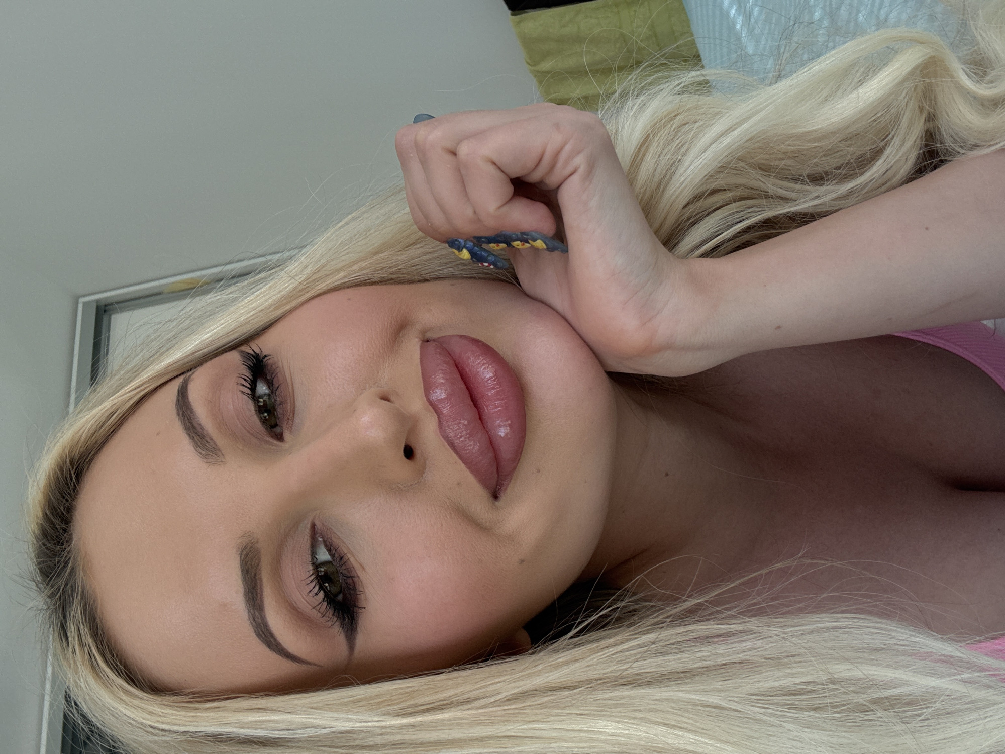 An overhead medium shot shows a fair-skinned blonde woman with long, wavy hair and full lips, looking at the camera with a slight smile. She has on dramatic makeup including heavy eyeliner, eyeshadow, and lip gloss. Her right hand is resting on her chin, and she has elaborate nail art on her fingernails. She is wearing a pink garment that is visible at the neckline. The background consists of a light gray wall and a sliver of a green object.