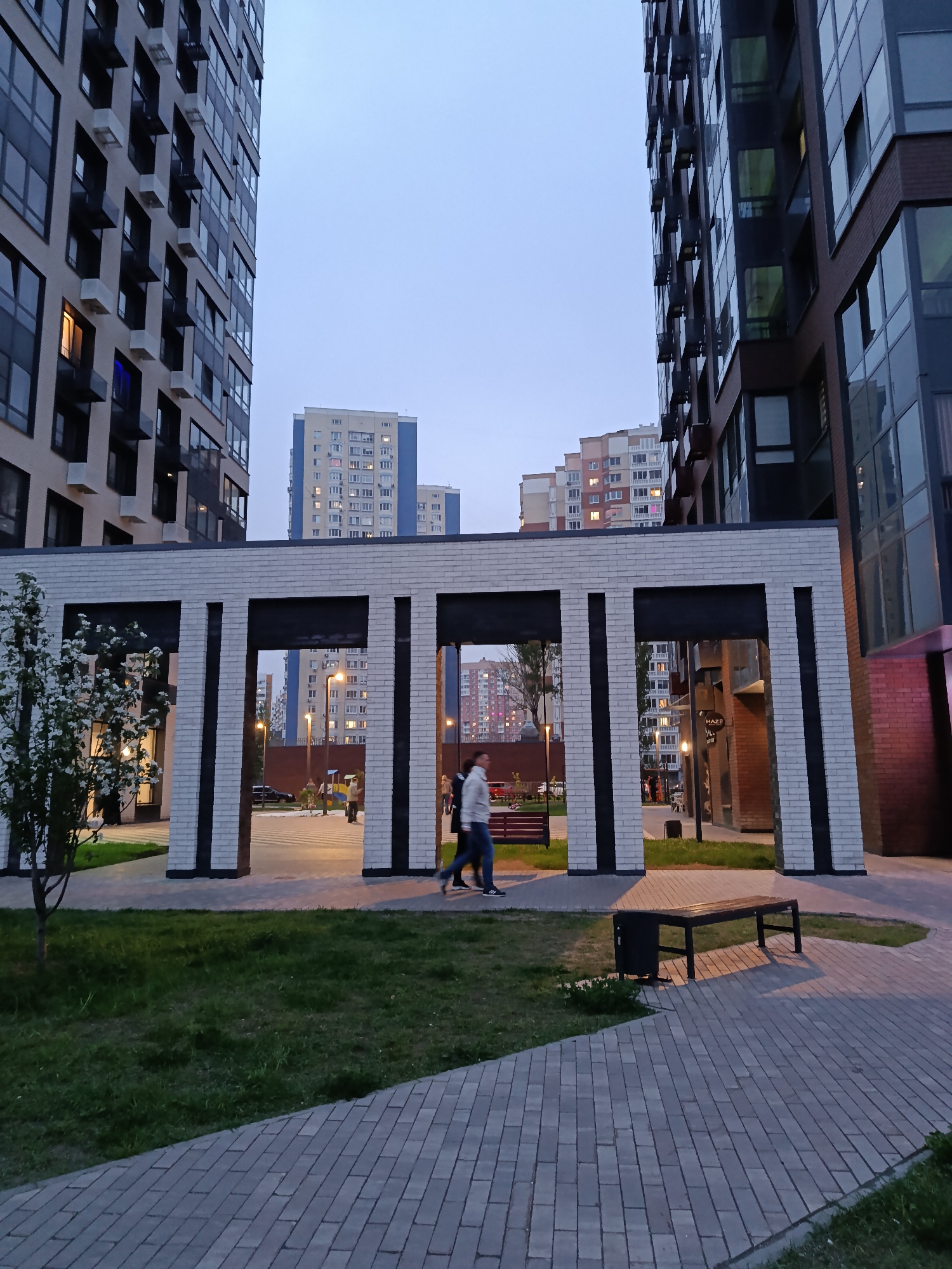 The image depicts a courtyard in a residential area in Moscow, Russia, at dusk. Two tall apartment buildings frame the scene, with other multi-story buildings visible in the background. A decorative, partially open structure of white brick with black accents stands in the middle ground, creating a gateway. Two people walk through this structure, appearing to be a man and a woman. A bench and a small trash receptacle are located on the right side of the image, along a paved walkway. A small tree with white blossoms is seen on the left. The courtyard features green grass and paved areas, and lighting suggests it is late in the day or early evening.