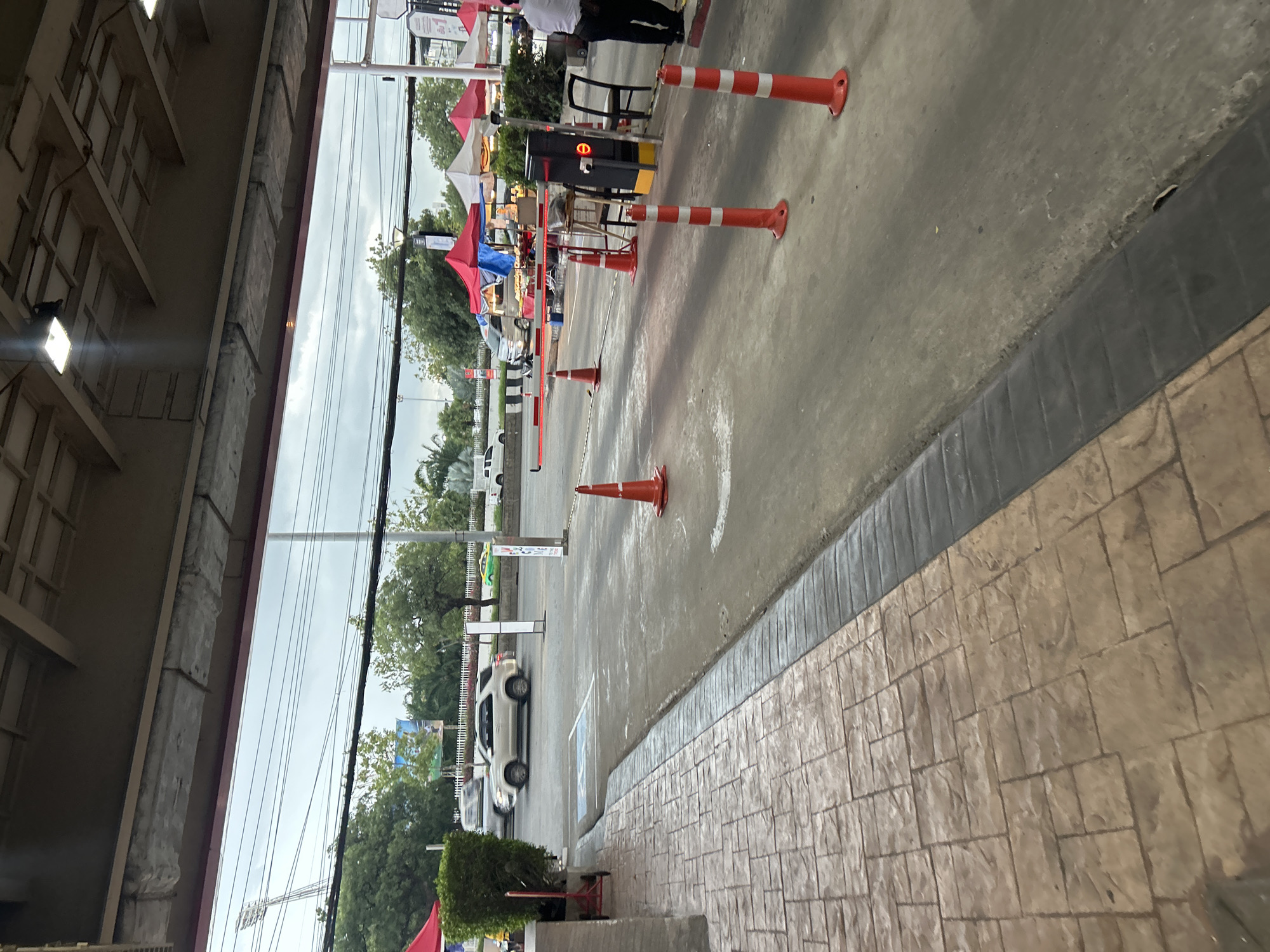 The image shows a view from under a covered walkway, looking out onto a street with a traffic barrier and several orange traffic cones. A silver SUV is driving on the road, and behind it, there are several market stalls with red and white canopies. Further in the background, there are trees and streetlights. The sky is overcast. The location is identified as Khlong Luang Town Municipality, Thailand. A man in a white shirt and dark pants is visible on the right side of the image, standing near the barrier.